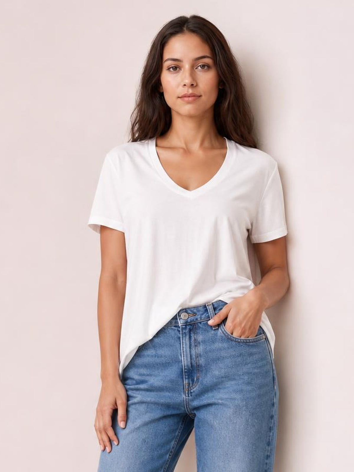 A model wears a white bamboo V-neck high-low T-shirt with short sleeves, a relaxed silhouette, and a slightly longer hem at the back while posing with one hand in her jeans pocket.