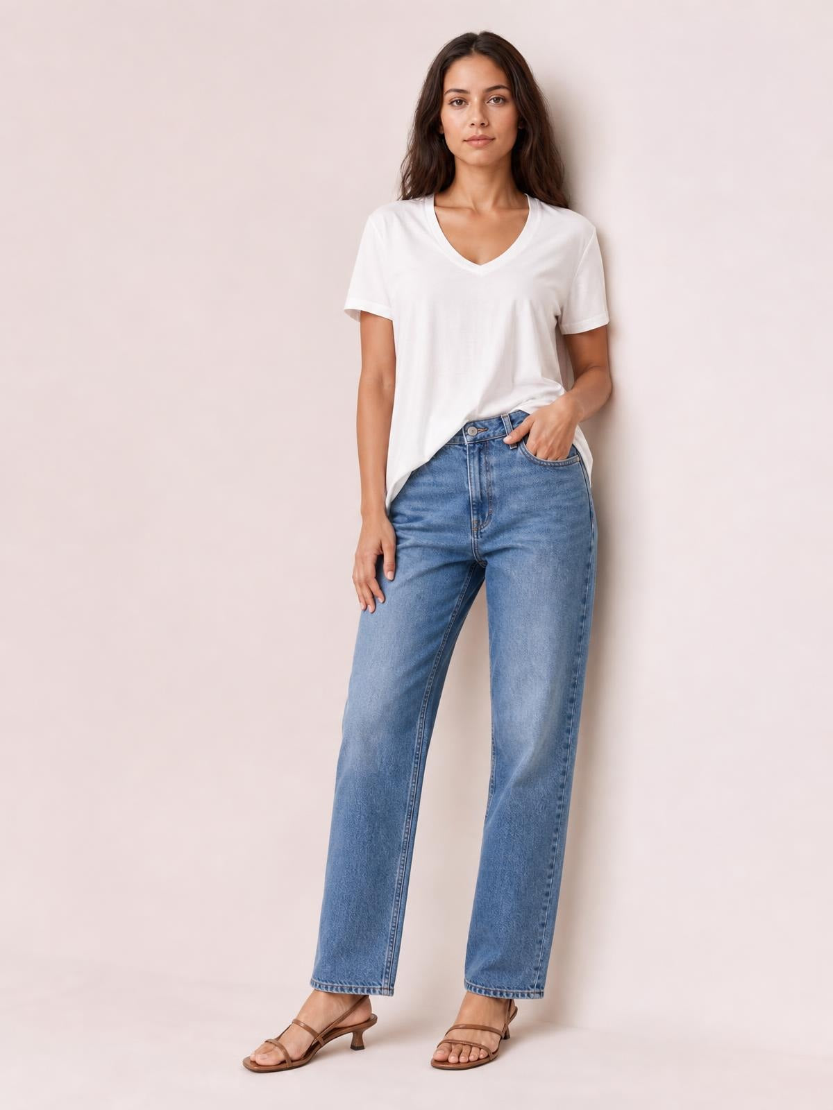 A model wears a white bamboo V-neck high-low T-shirt with short sleeves, a relaxed silhouette, and an uneven hem, posing with one hand in her jeans pocket.