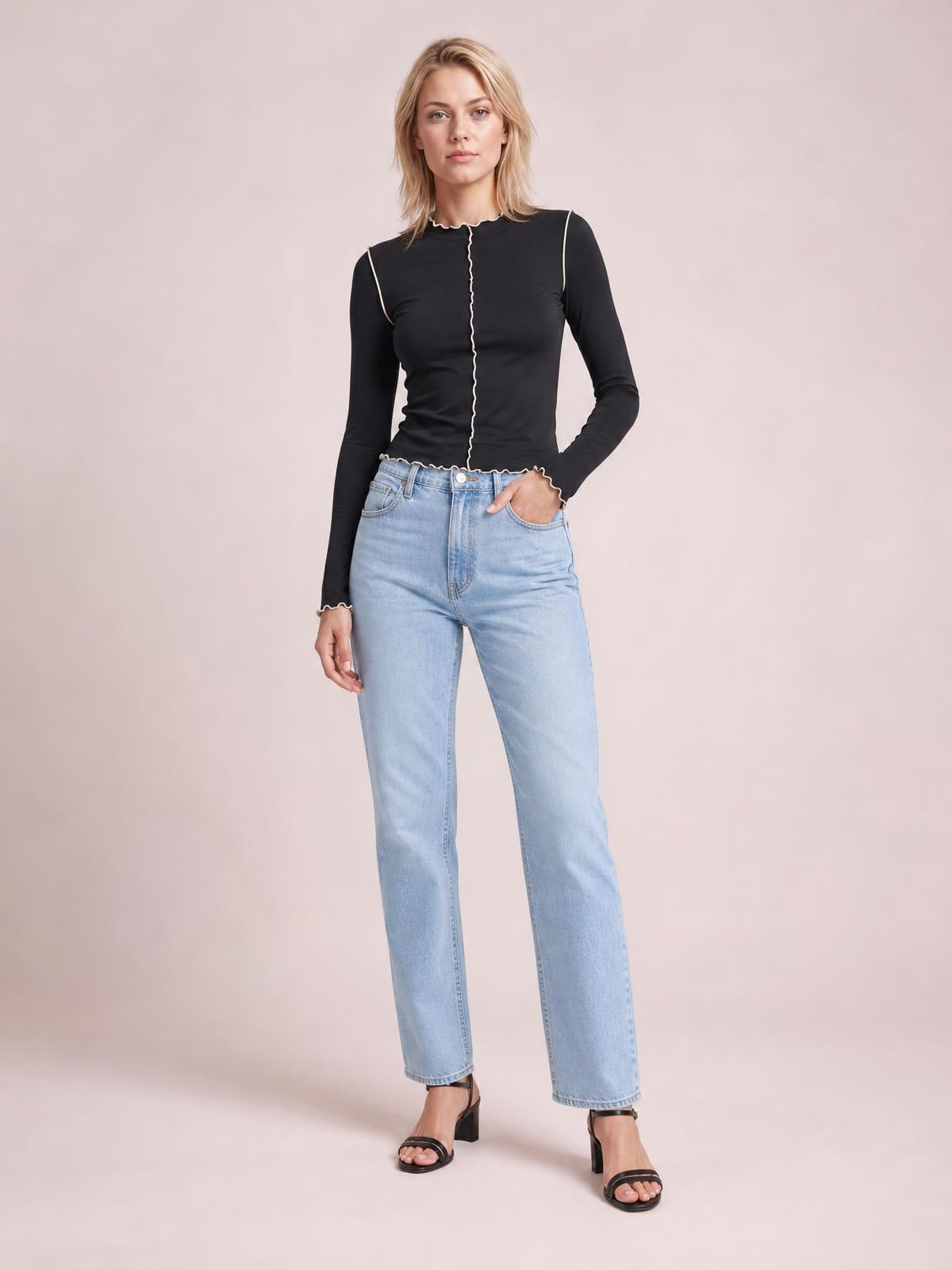A model wears a fitted black mock neck long sleeve T-shirt with contrasting white trim, a stretchy silhouette, and scalloped edges, posing with one hand in her jeans pocket.