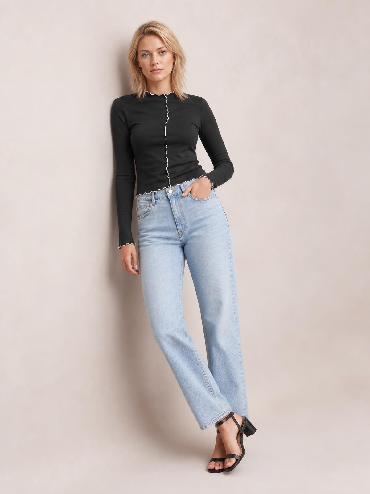 Model wears a fitted black mock neck long sleeve T-shirt with white contrast trim along the neckline, front seam, cuffs, and hem, posing with one hand in her jeans pocket.