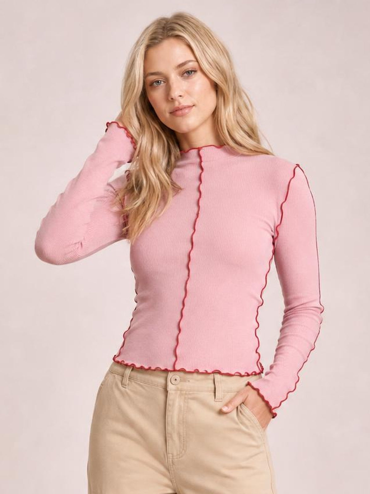 Model poses in a pink fitted mock neck long sleeve T-shirt with contrasting red trim along the neckline, seams, cuffs, and hem.