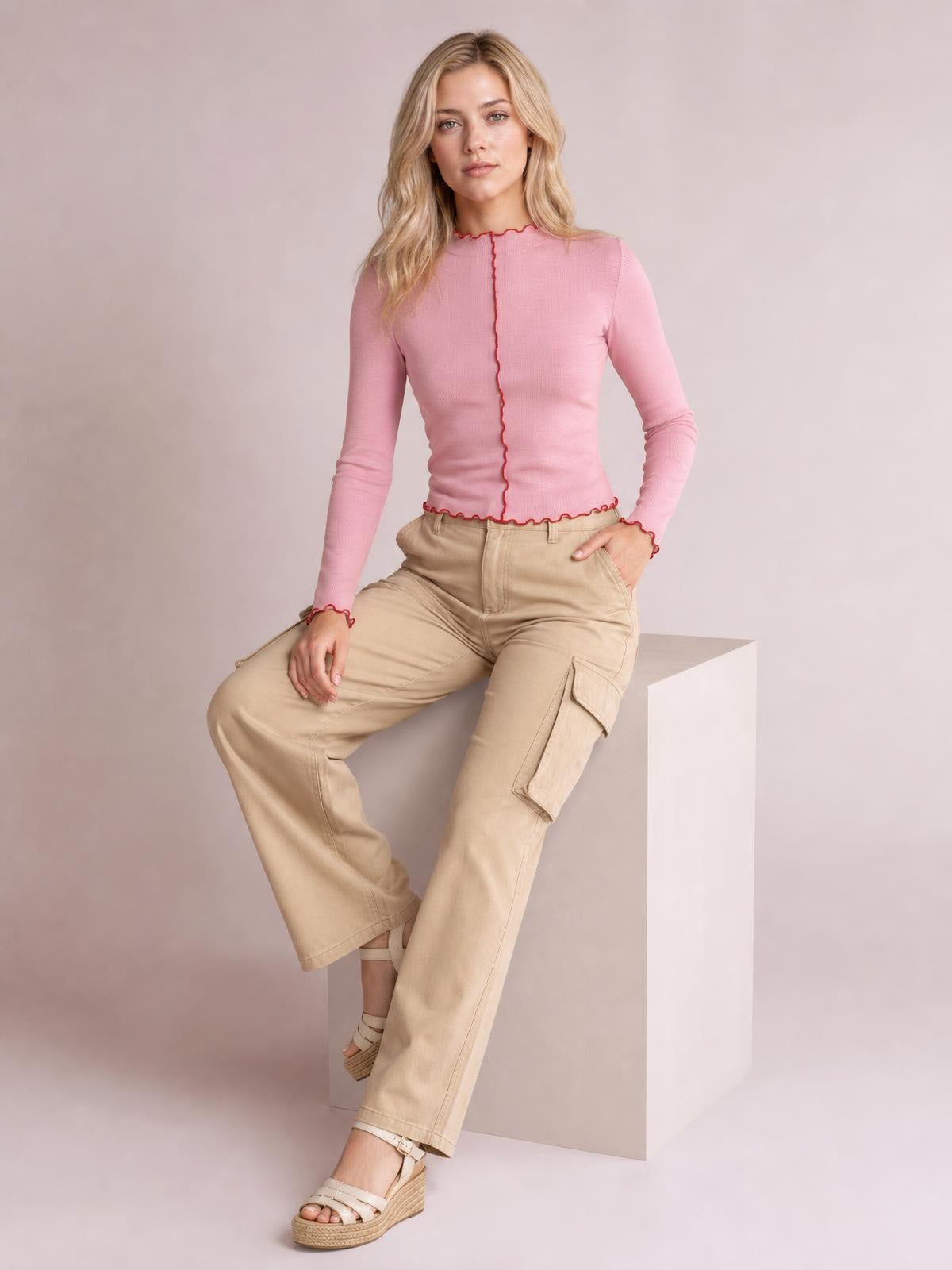 A model sits in a fitted pink mock neck long sleeve T-shirt with red contrast trim along the collar, center seam, cuffs, and hem.
