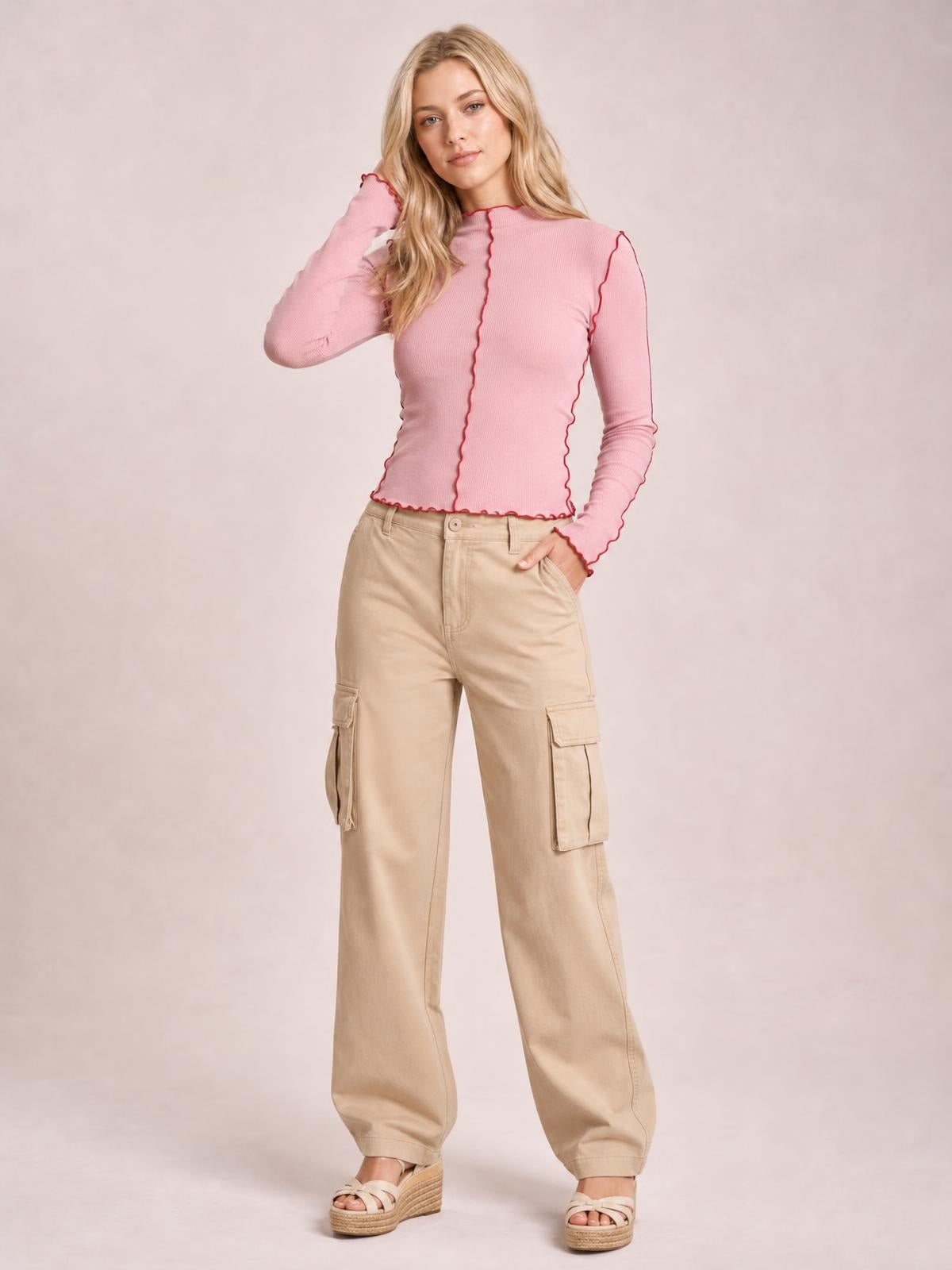 Model poses with one hand in her pocket, wearing a fitted pink mock neck long sleeve T-shirt with red contrast trim and scalloped edges.