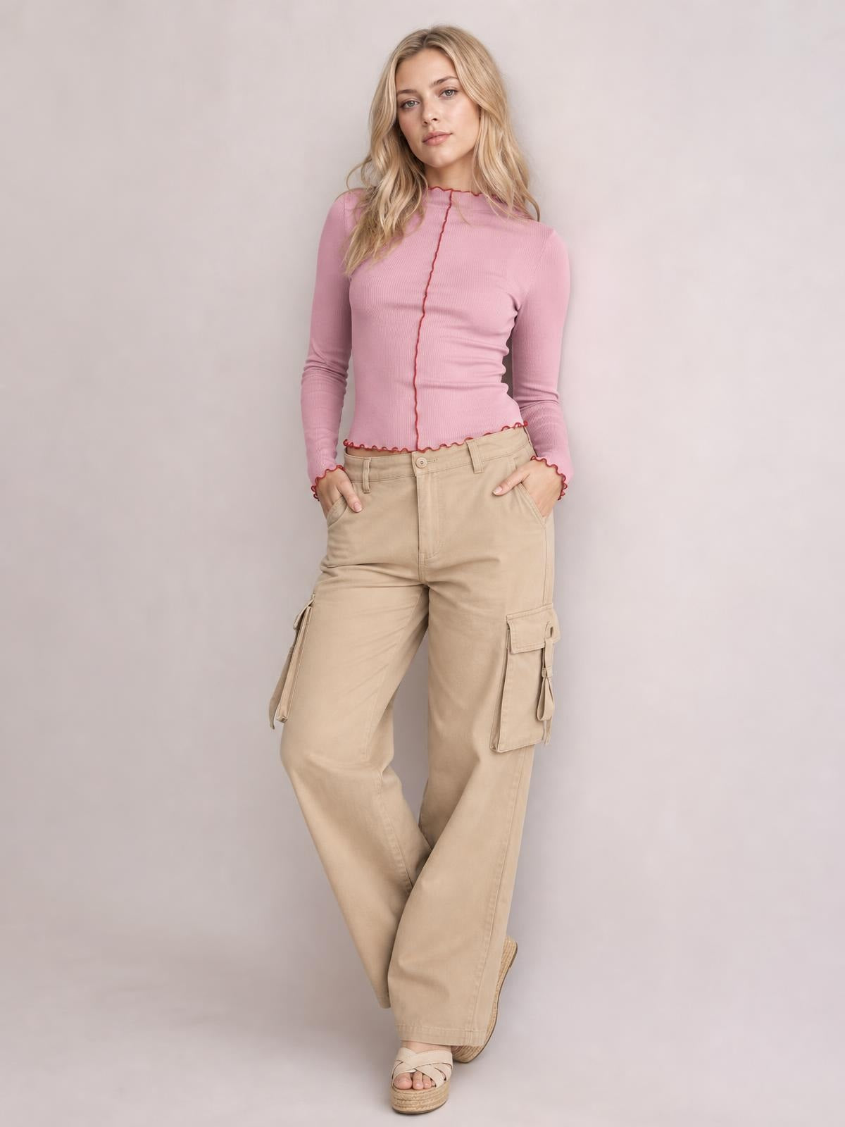 A model poses with hands in her pockets wearing a pink fitted mock neck long sleeve T-shirt with red contrast trim along the neckline, center seam, and hems.