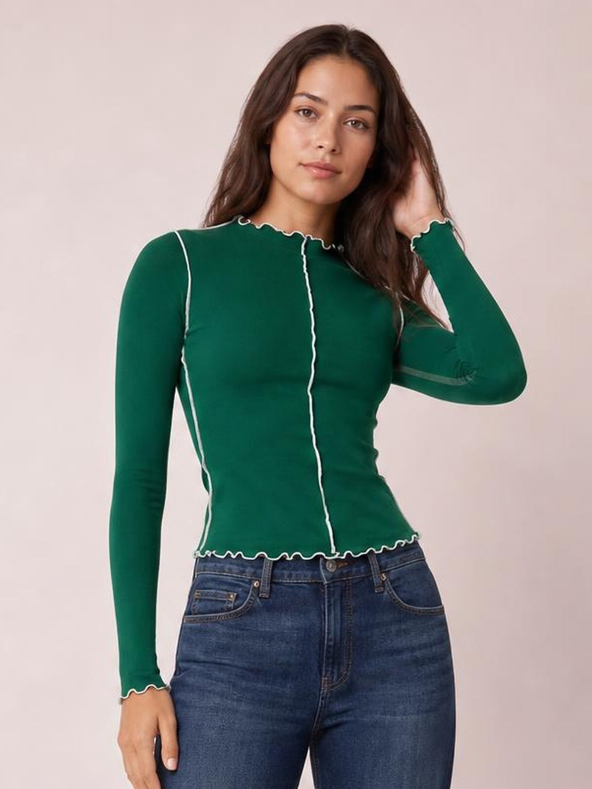 Model wearing a fitted green mock neck long sleeve T-shirt with white contrast trim, scalloped edges, and vertical seam detailing while touching her hair.