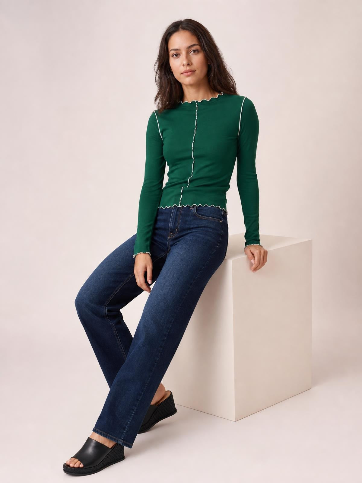 Model seated in a fitted dark green mock neck long sleeve T-shirt with white contrast trim along the shoulders, center seam, neckline, cuffs, and hem.