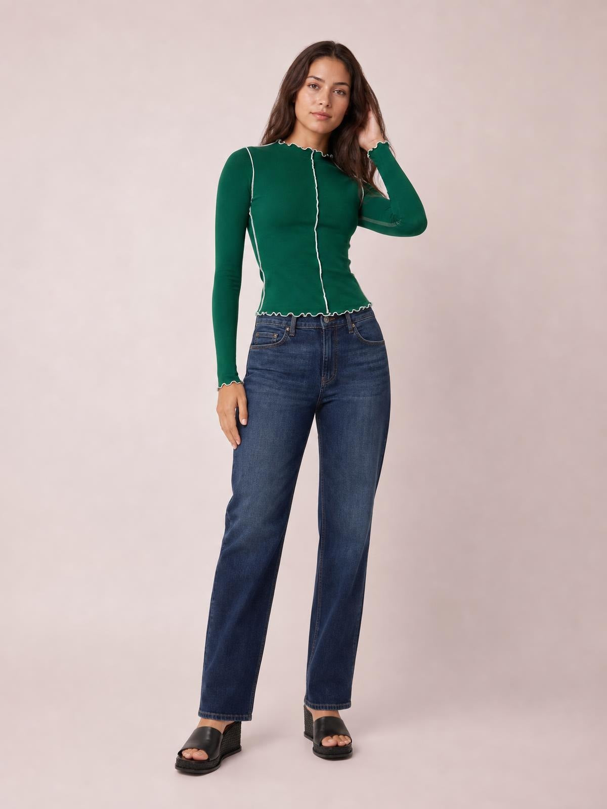 Model in a fitted forest green mock neck long sleeve T-shirt with white contrast trim, scalloped edges, and a slim silhouette, posing with one hand in her hair.