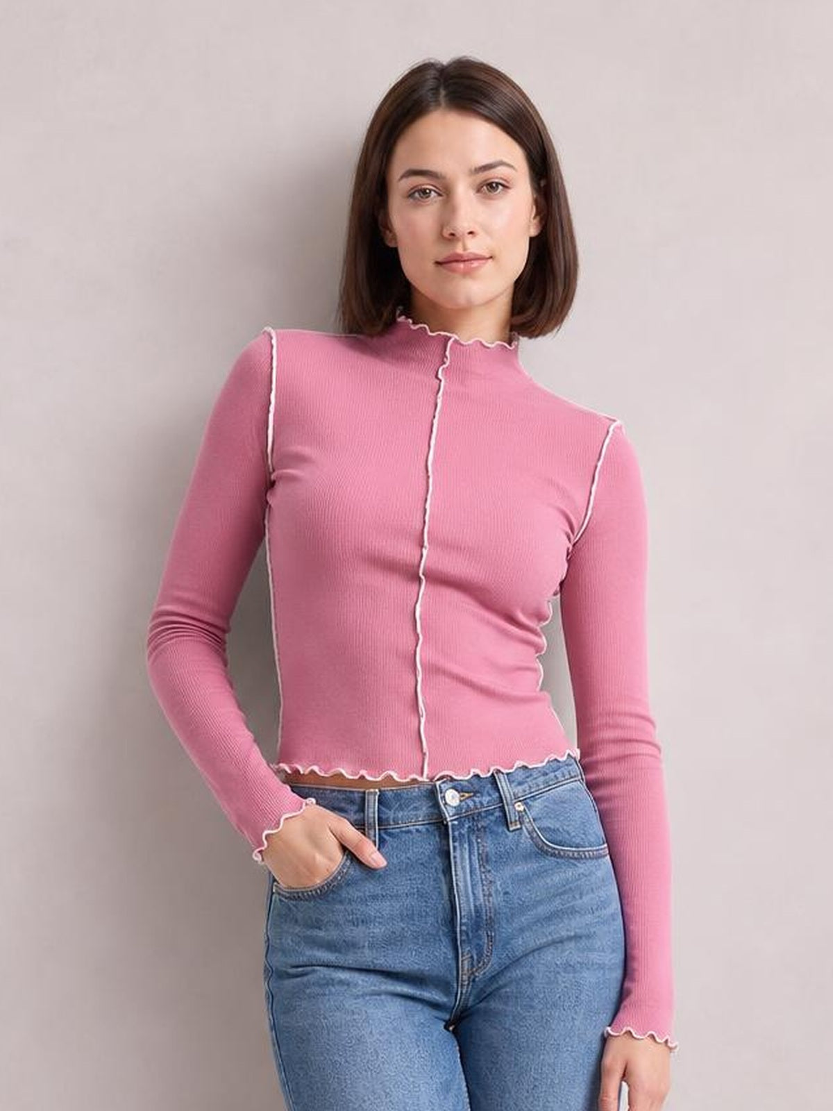 A model poses in a fitted pink contrast trim mock neck long sleeve T-shirt with white edging, ribbed construction, and a scalloped hem and cuffs.