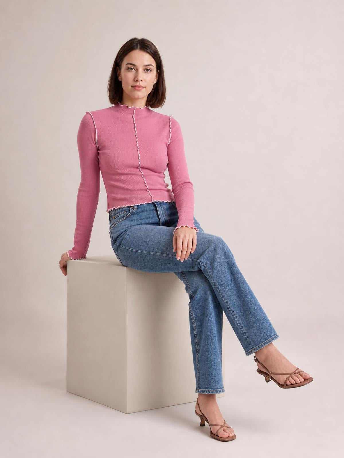 A seated model wears a fitted pink mock neck long sleeve T-shirt with contrasting white trim, stretchy silhouette, and scalloped edges.