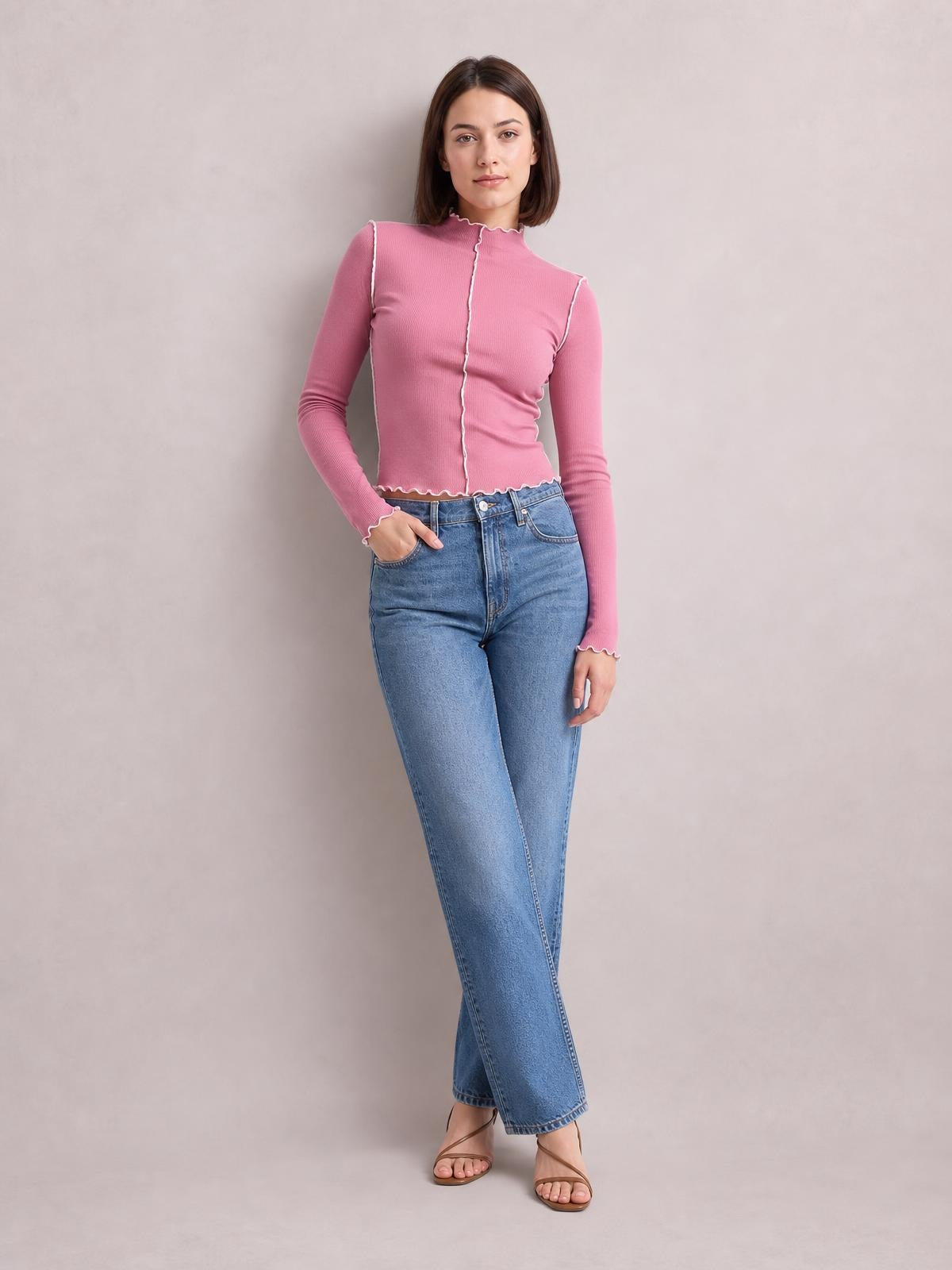 Model stands with one hand in her jeans pocket wearing a fitted pink mock neck long sleeve T-shirt with contrasting white trim and scalloped edges.
