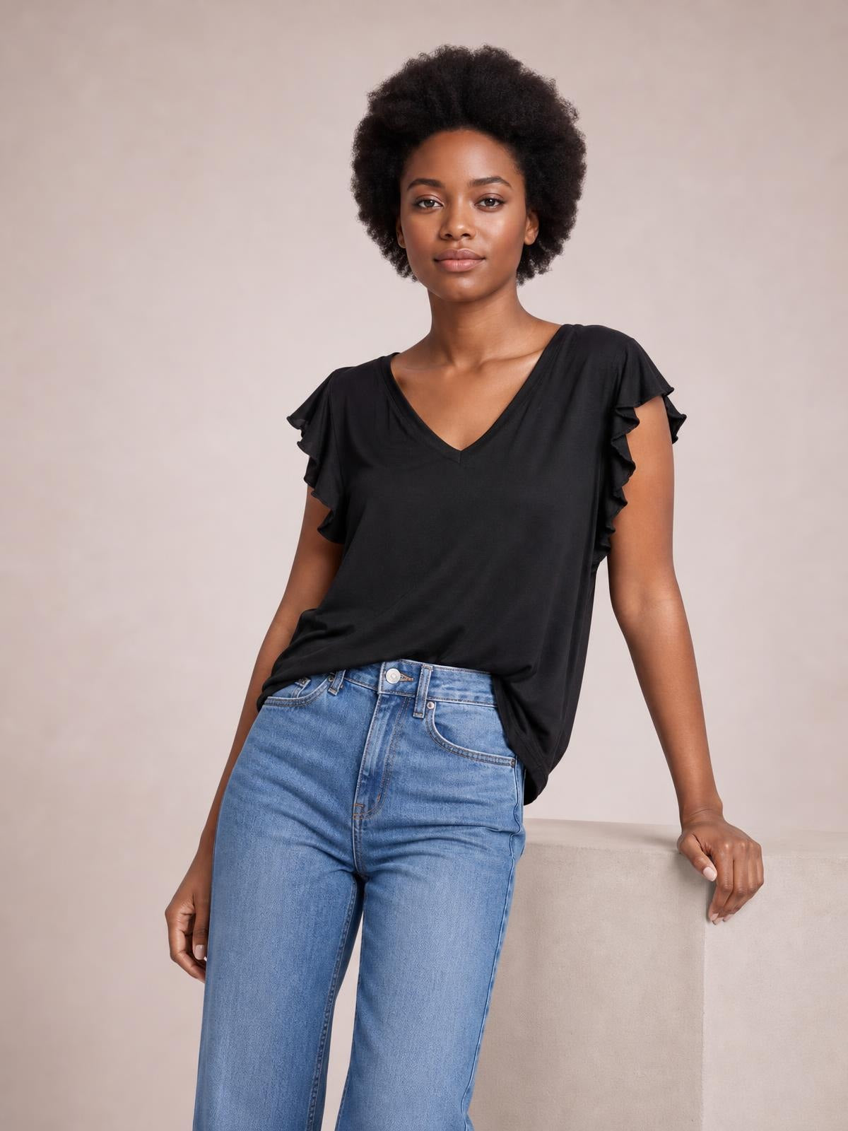 Model poses beside a chair wearing a black bamboo T-shirt with a notched V-neck, relaxed silhouette, and layered ruffled short sleeves.
