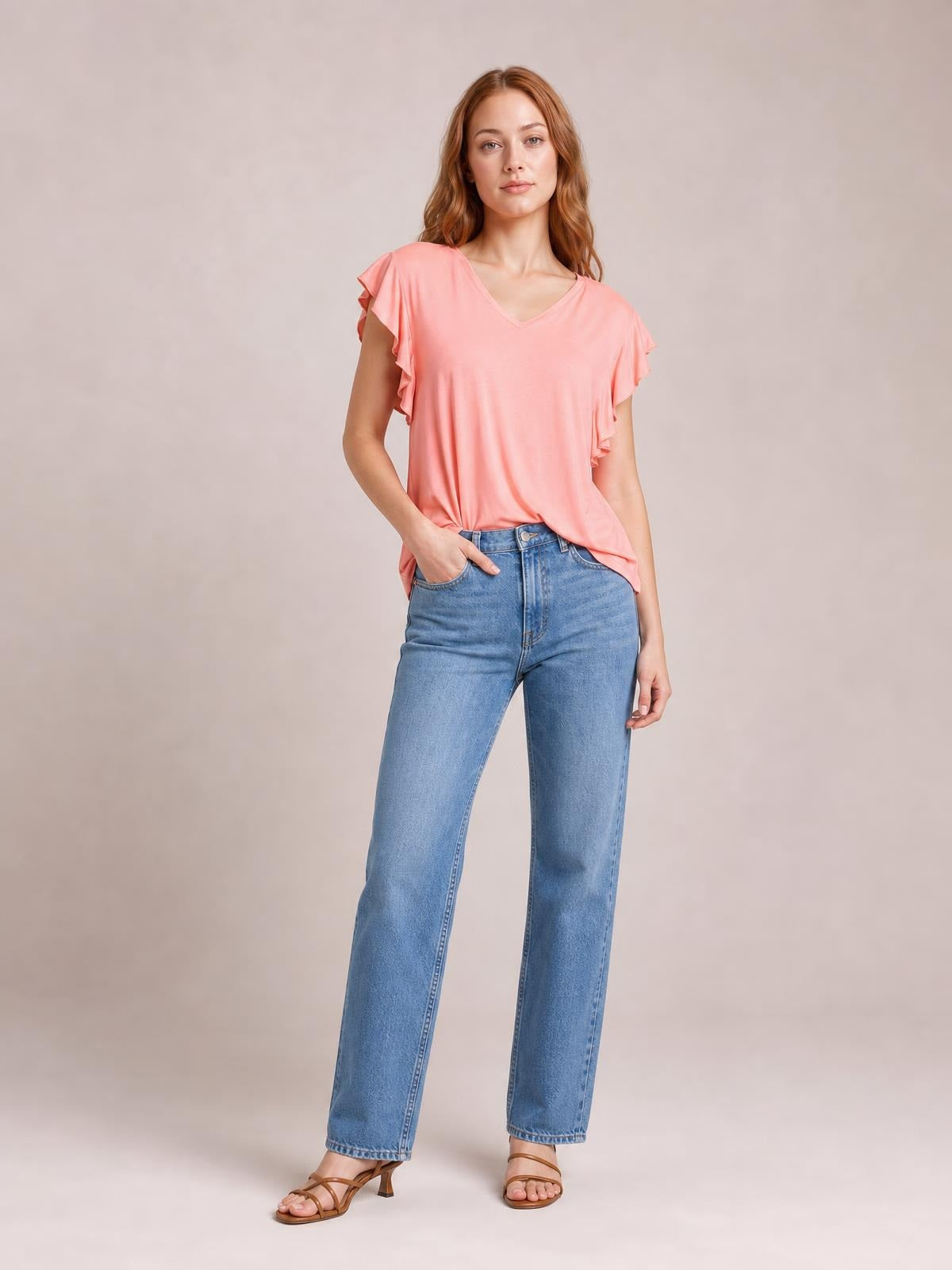 Model wearing a coral bamboo T-shirt with a notched V-neck, ruffled short sleeves, and a relaxed, stretchy silhouette, posing with one hand in her jeans pocket.