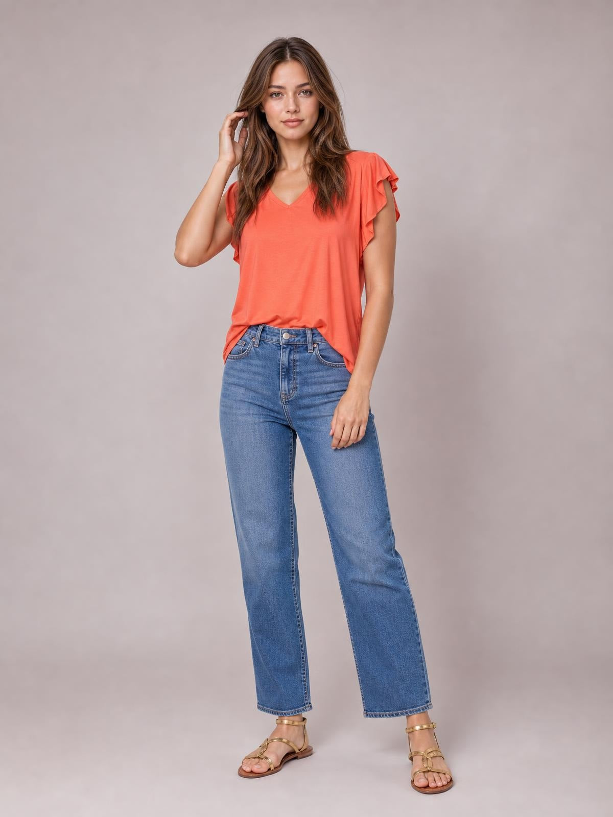 A model poses in a coral bamboo T-shirt with a notched V-neck, ruffled short sleeves, and a relaxed silhouette, one hand touching her hair.