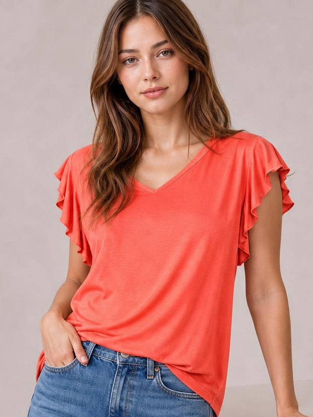 Model wearing a coral bamboo T-shirt with a notched neckline, ruffled short sleeves, and a relaxed silhouette, standing with one hand in her jeans pocket.