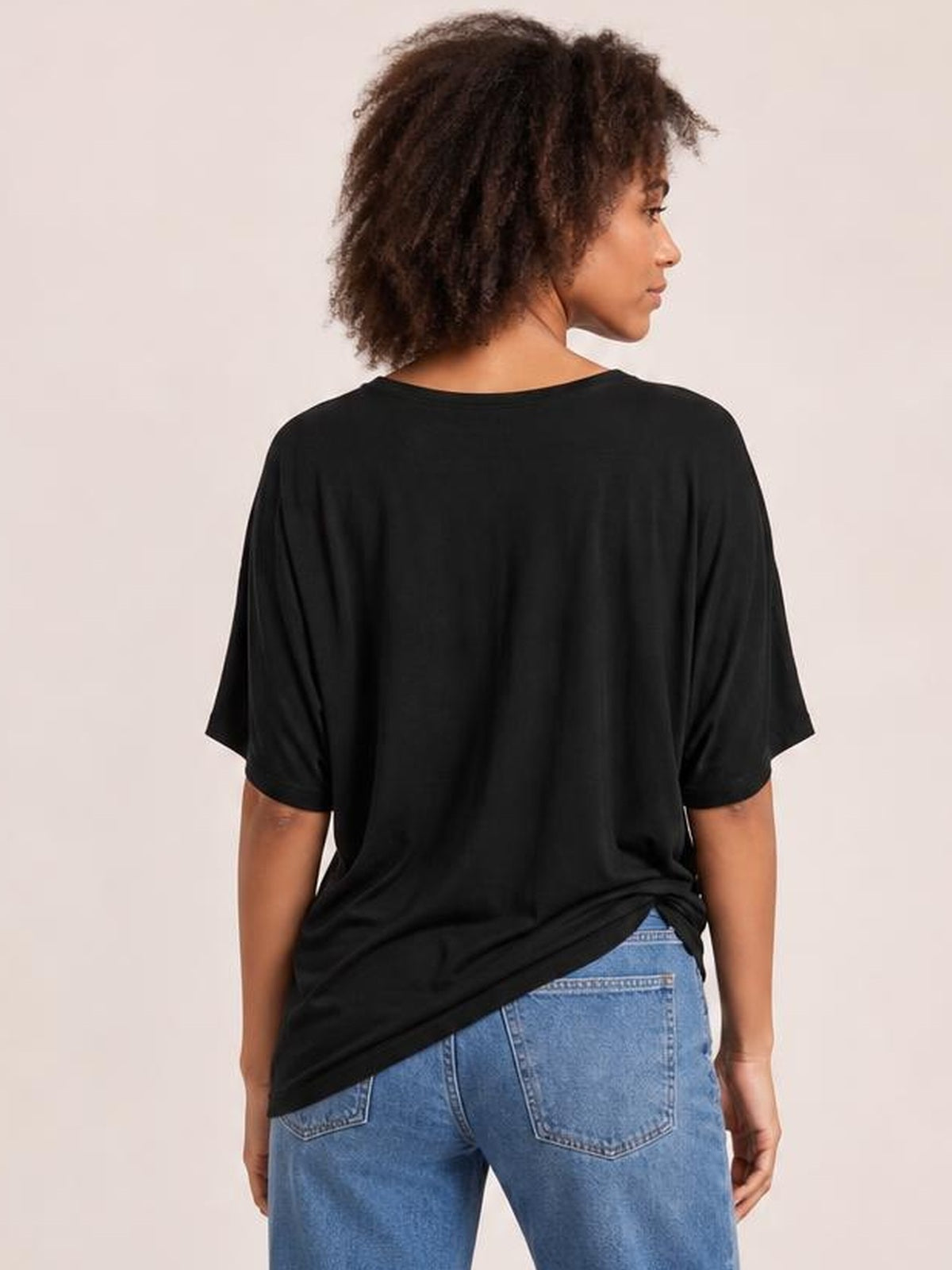 A model looks over her shoulder while wearing a black drop-shoulder T-shirt with a loose silhouette, short sleeves, and a longer hem over blue jeans.