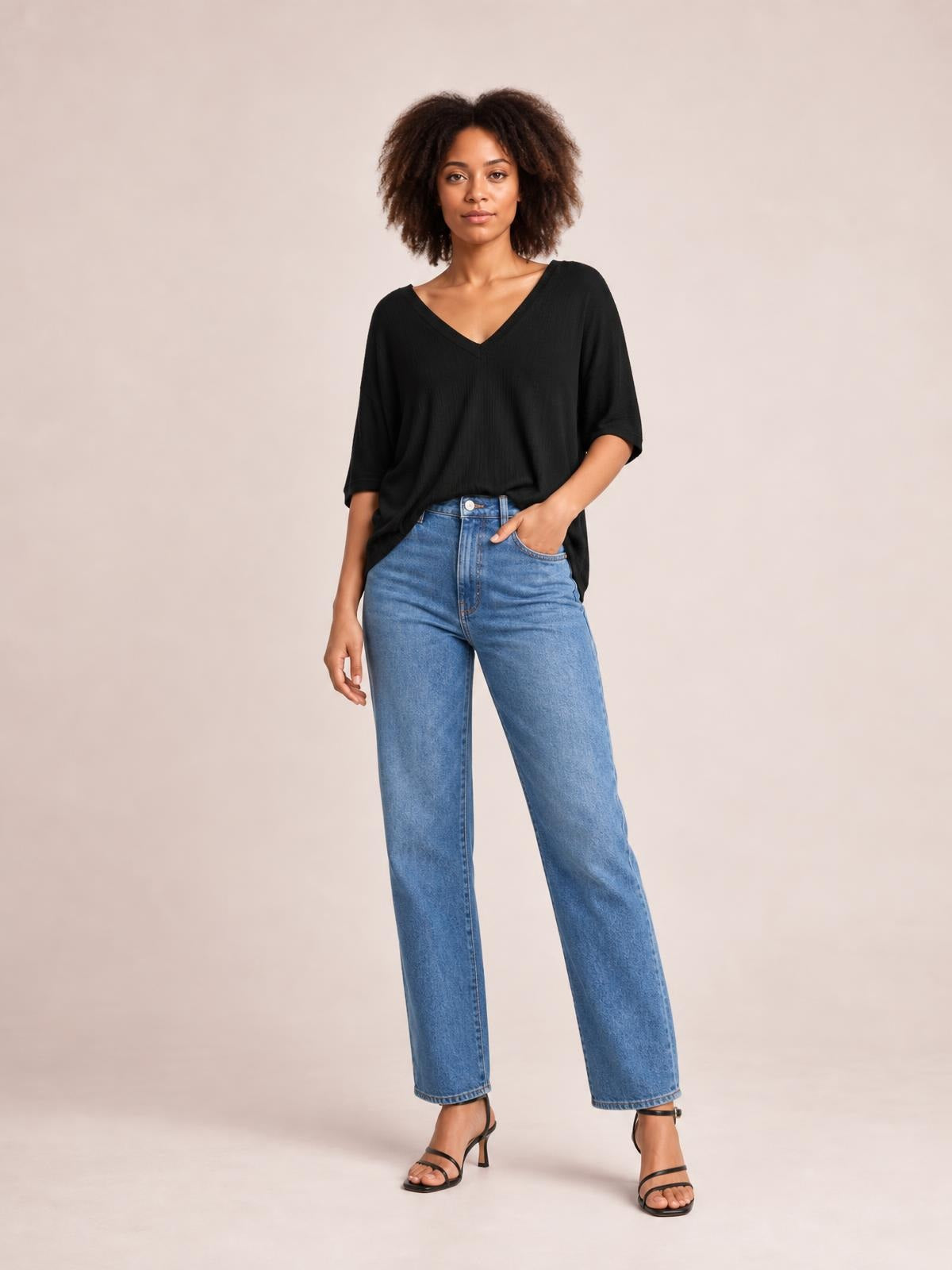 Model wearing a black bamboo V-neck drop shoulder T-shirt with a relaxed silhouette and elbow-length sleeves, posed with one hand in her jeans pocket.