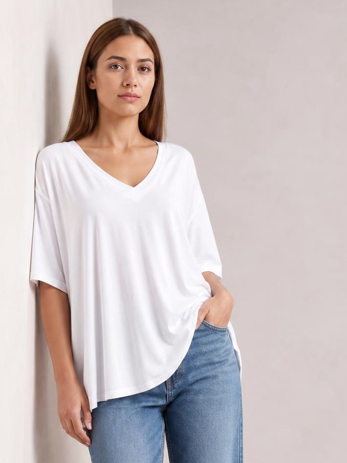 Model wearing a white Bamboo V-neck drop shoulder T-shirt with a relaxed, oversized silhouette, short sleeves, curved hem, and one hand in her jeans pocket.