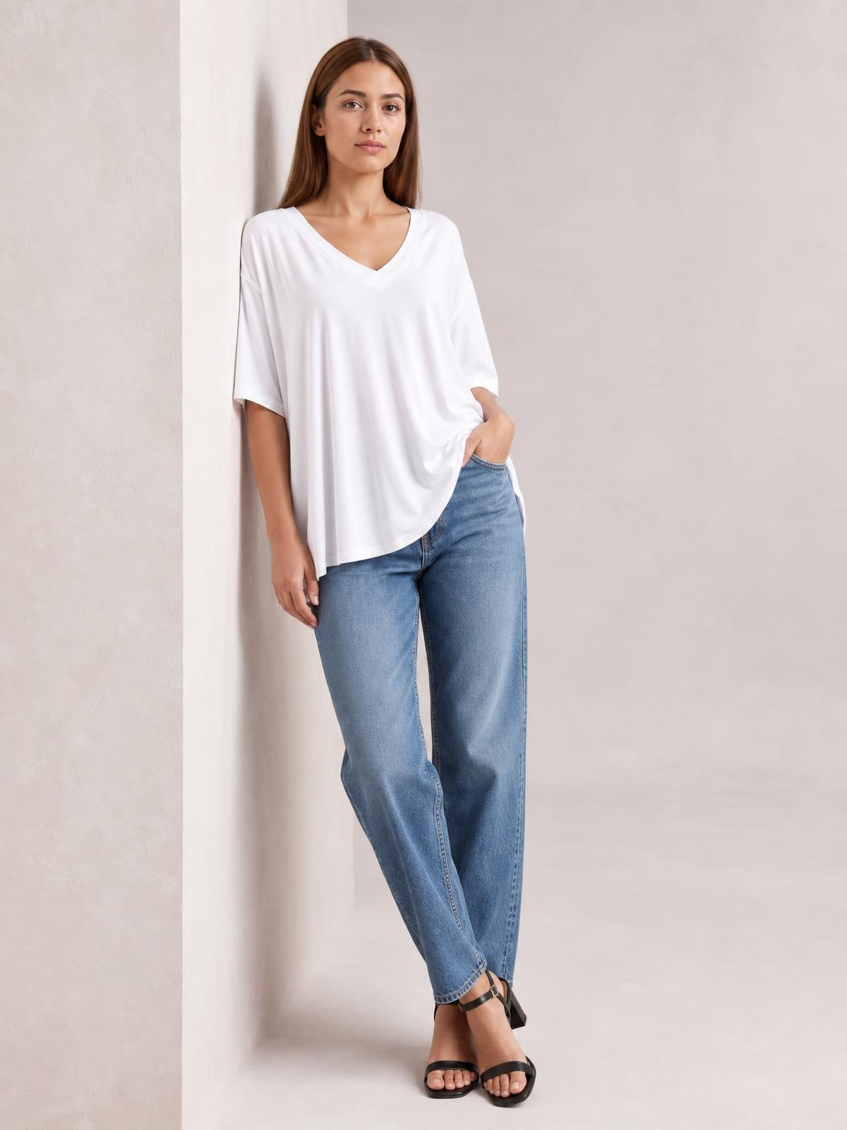 A woman poses in a white bamboo V-neck drop shoulder T-shirt with a relaxed, oversized silhouette and short sleeves, resting one hand in her jeans pocket.