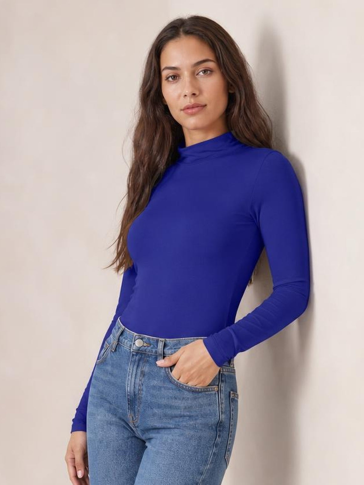Model wears a cobalt blue mock neck long sleeve bodysuit with a fitted silhouette and smooth opaque construction, paired with high-waisted jeans.