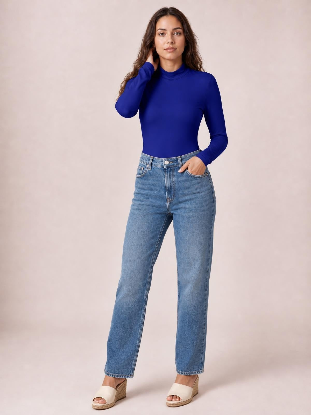 Model wears a royal blue mock neck long sleeve bodysuit with an opaque, fitted silhouette and smooth construction, posing with one hand at her neck.
