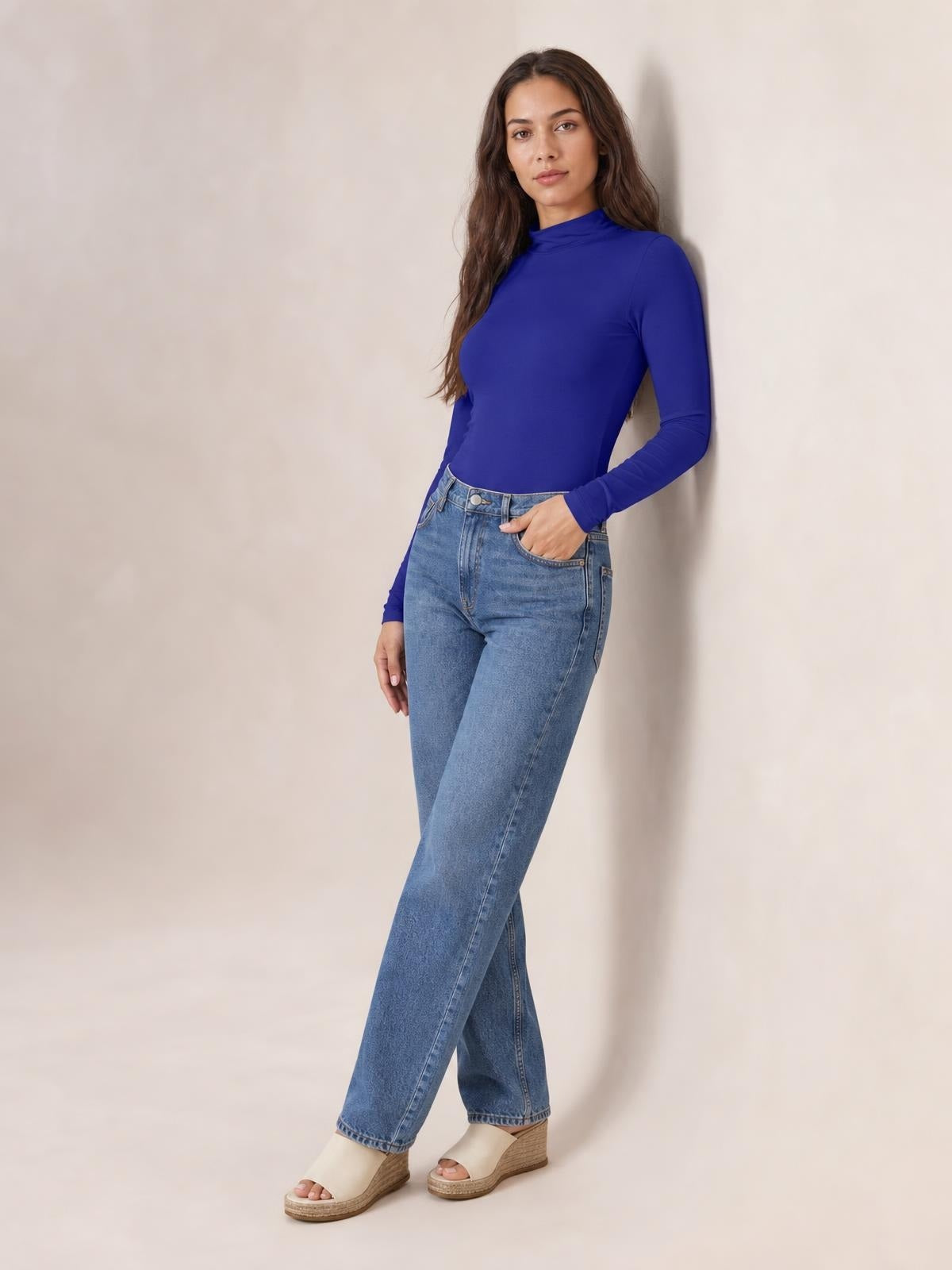 Model poses in a cobalt blue mock neck long sleeve bodysuit with a fitted silhouette and smooth opaque stretch construction, paired with jeans.