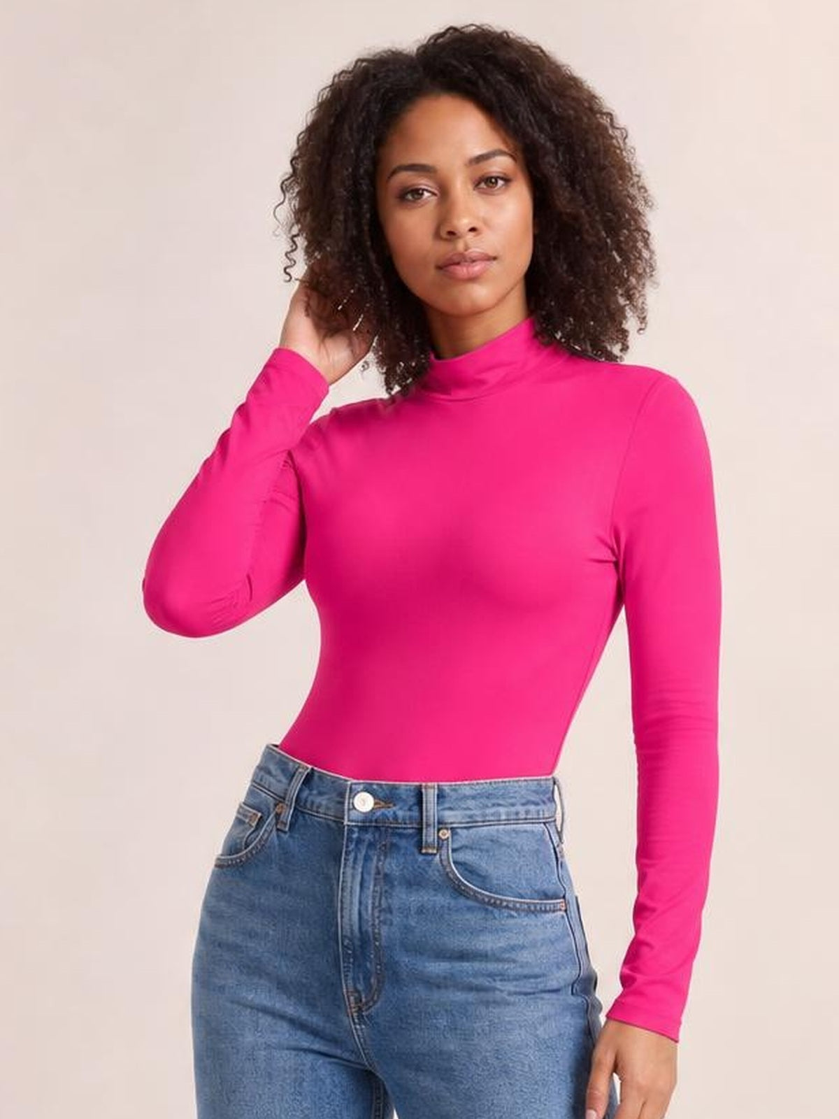Model poses in a vibrant pink mock neck long sleeve bodysuit with a fitted silhouette and smooth opaque construction, paired with jeans.