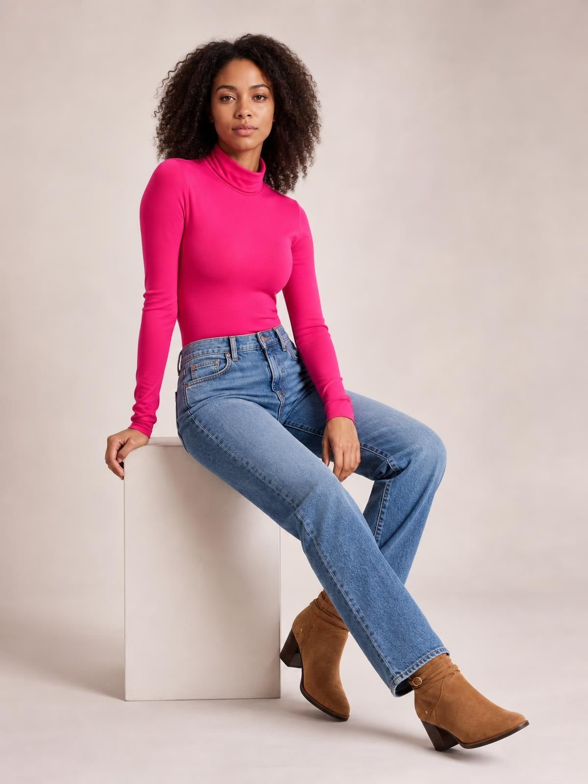 Model sits in a vibrant pink mock neck long sleeve bodysuit with a fitted silhouette, smooth opaque construction, and moderate stretch.