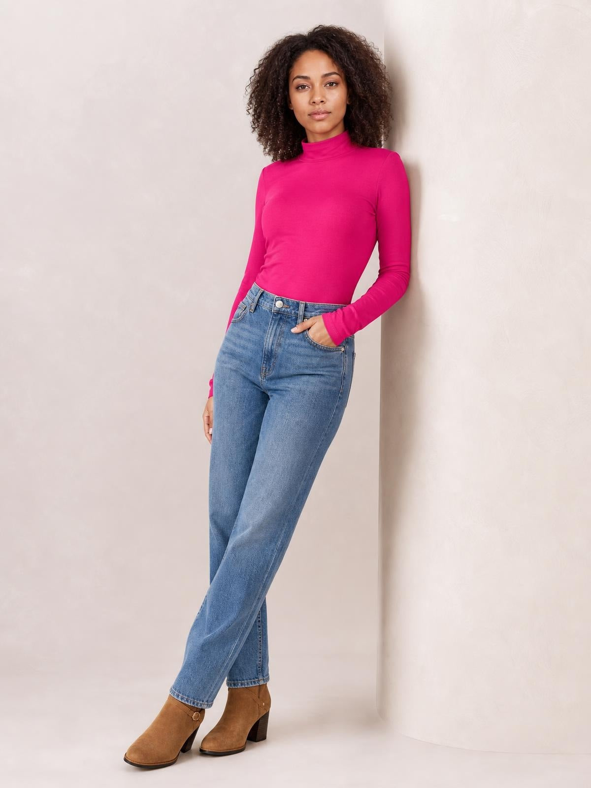 A model wears a bright pink mock neck long sleeve bodysuit with a fitted silhouette, tucked into jeans while leaning with one hand in a pocket.