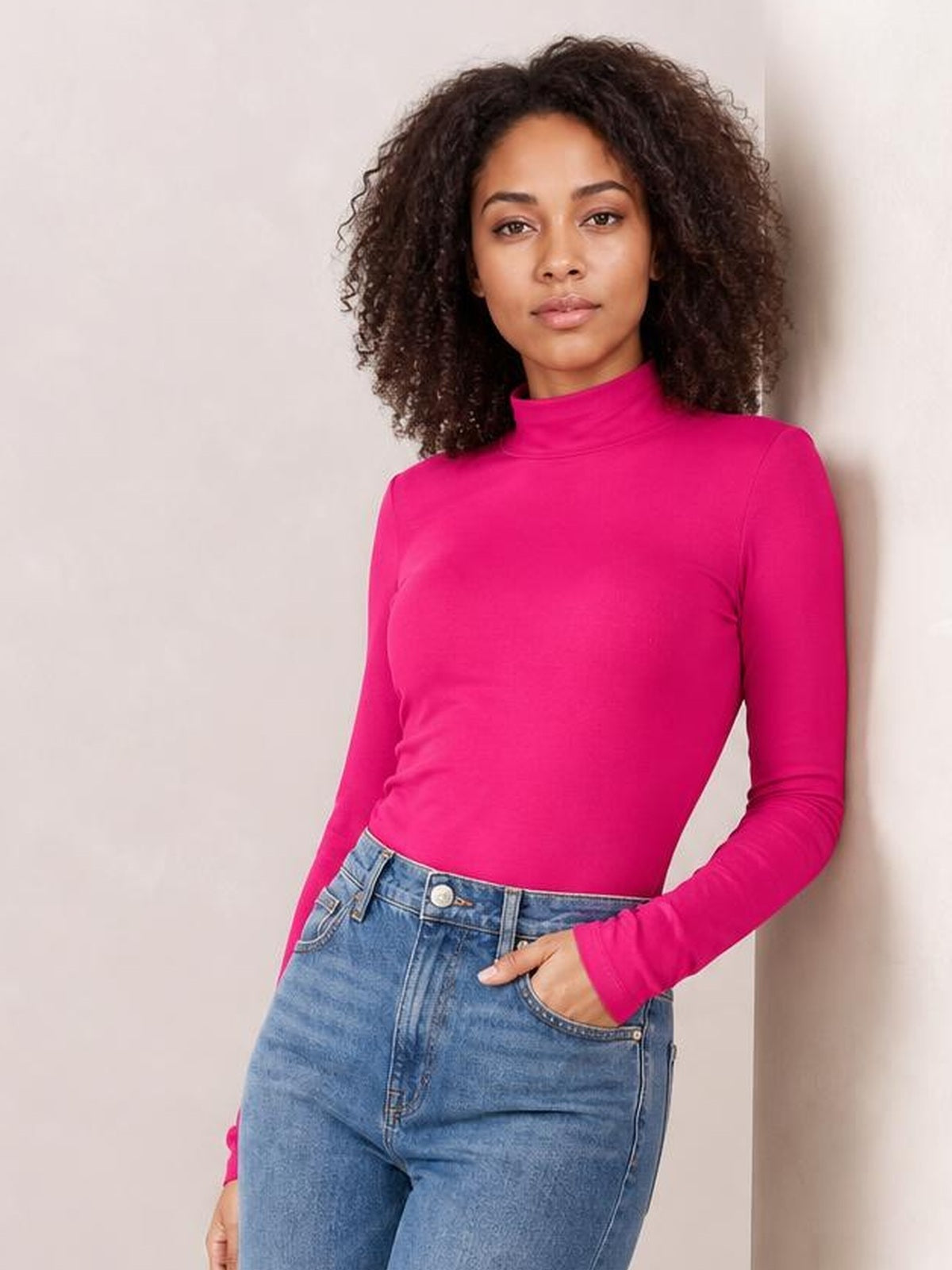 Model wearing a bright pink mock neck long sleeve bodysuit with an opaque, fitted silhouette and moderate stretch, leaning with one hand in her jeans pocket.