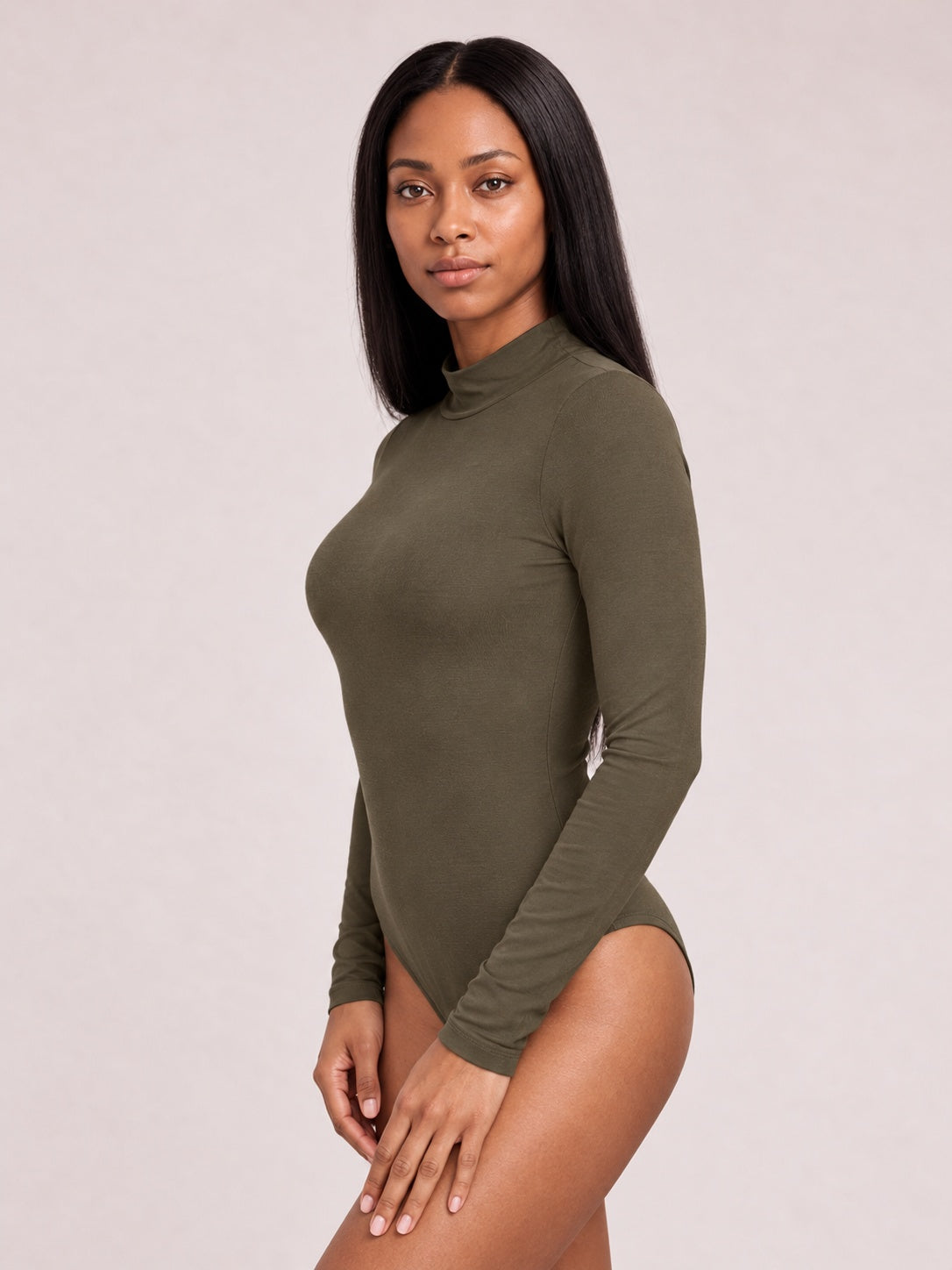Model poses in a three-quarter stance wearing an olive green mock neck long sleeve bodysuit with a fitted silhouette and high-cut bottom.