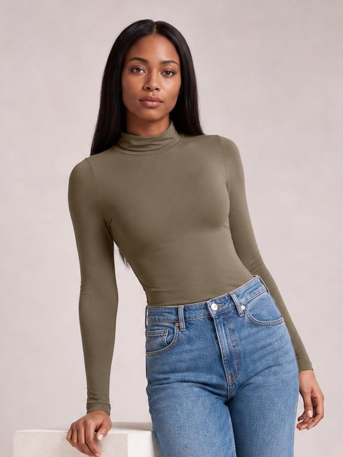 Model leans against a ledge wearing an olive mock neck long sleeve bodysuit with a smooth, fitted silhouette and full-length sleeves.