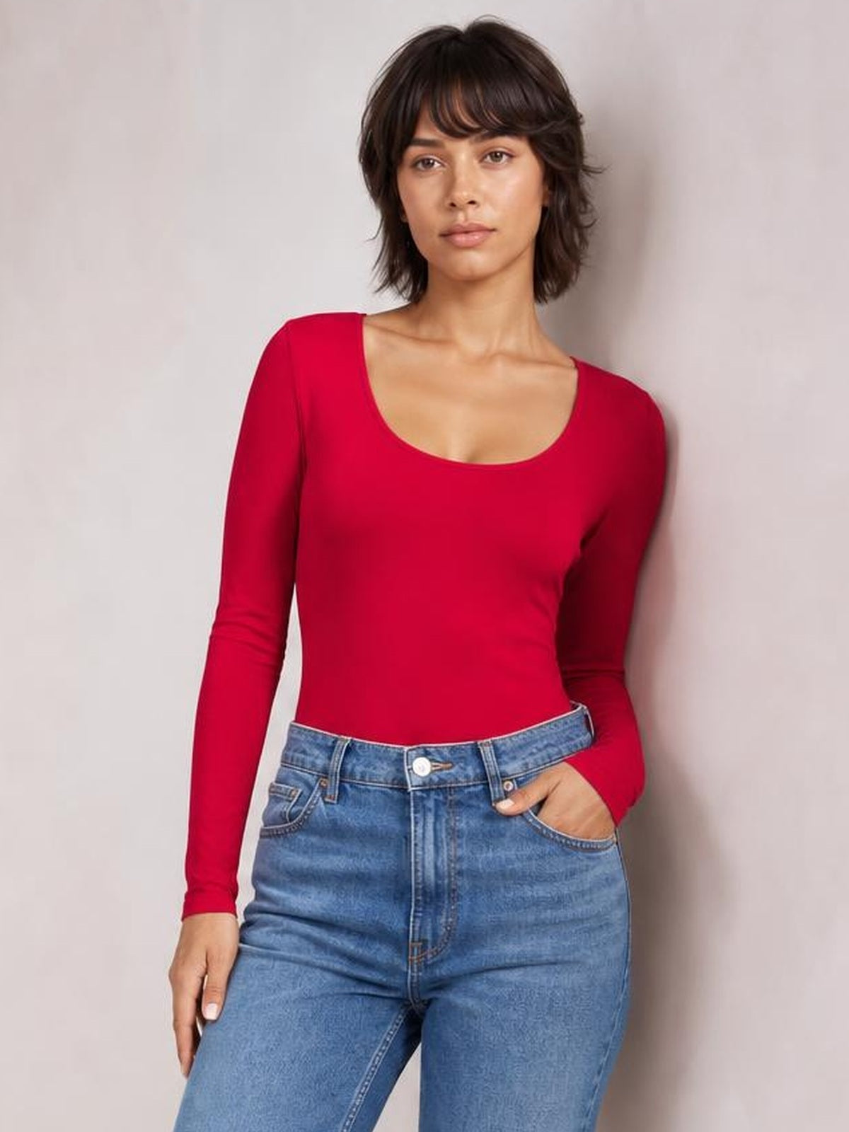 Model wears a red long-sleeve bodysuit with a scoop neckline and fitted silhouette, posing with one hand in her jeans pocket.