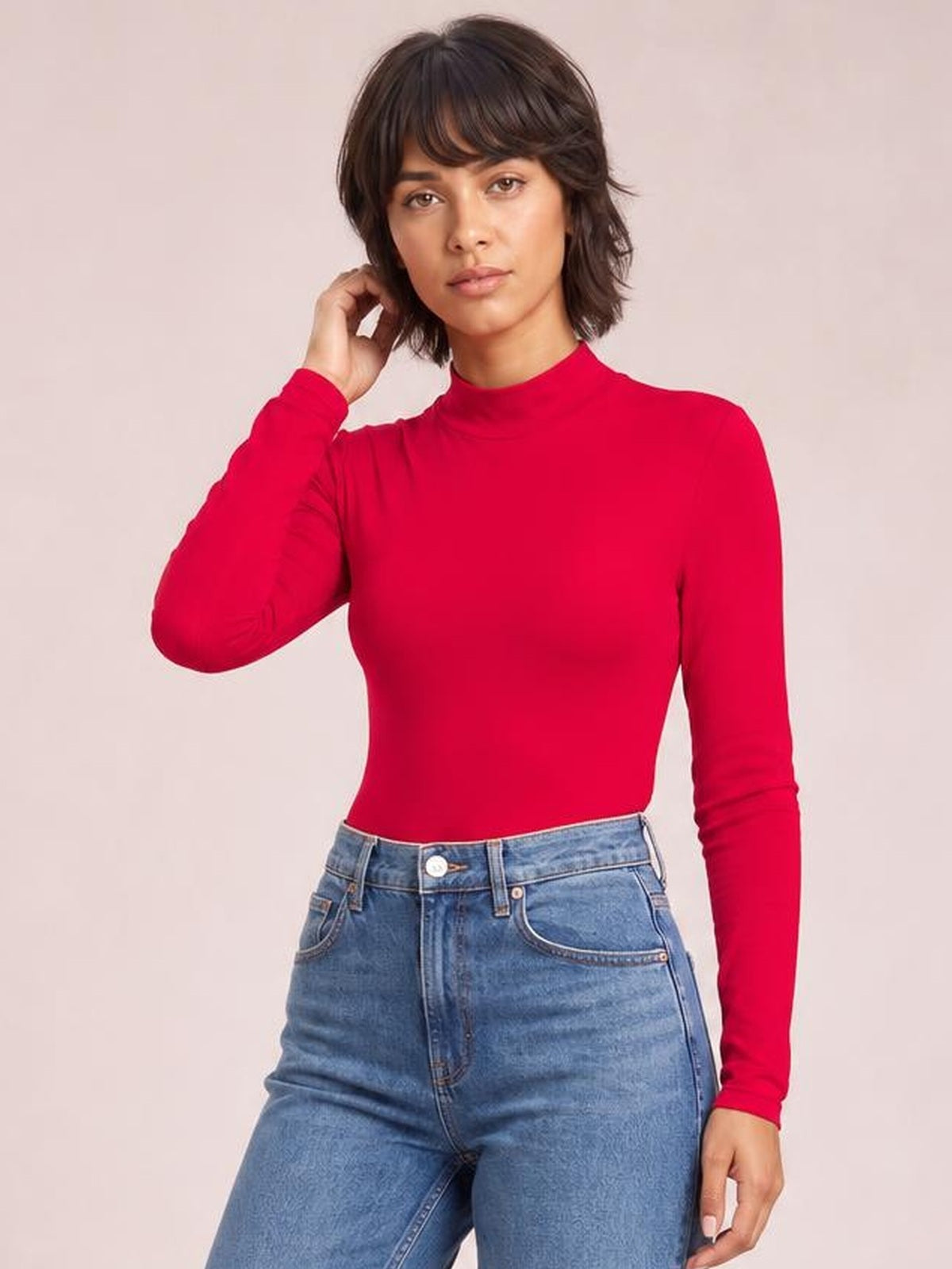 Model wears a bright red mock neck long sleeve bodysuit with a fitted silhouette, posing with one hand near her hair and tucked into blue jeans.
