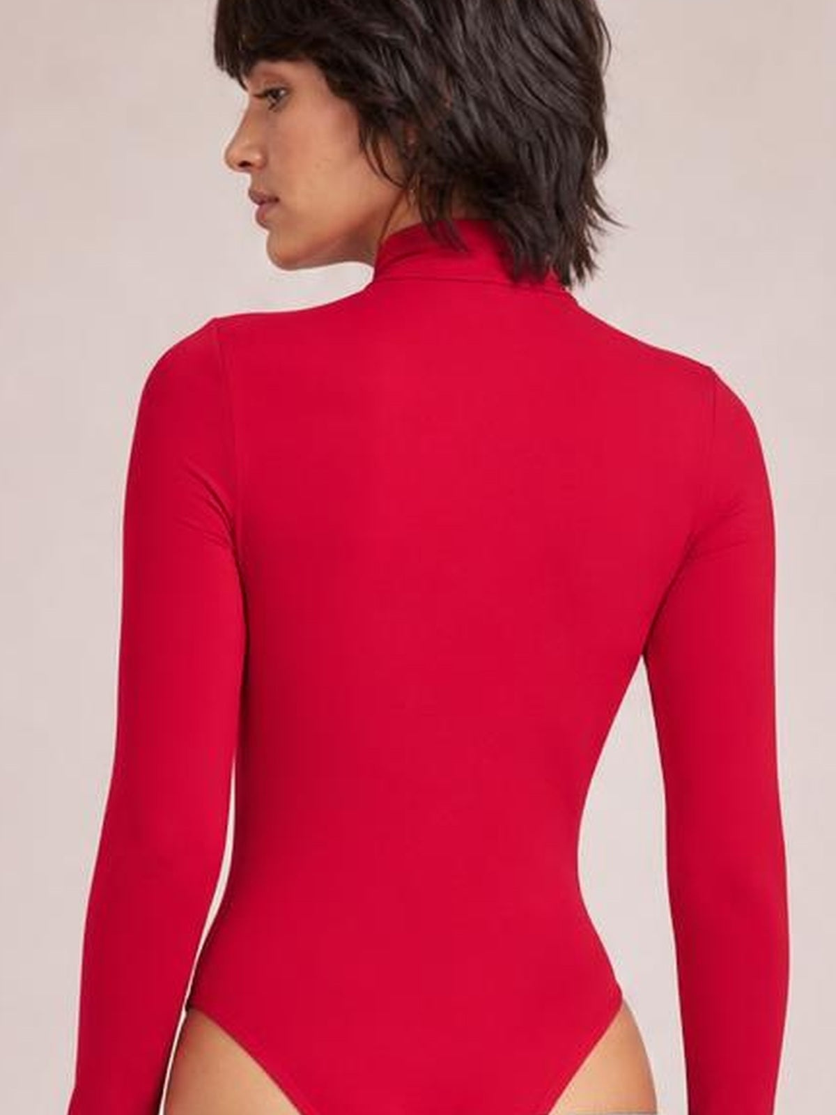 Model turned away in a bright red mock neck long sleeve bodysuit with a fitted silhouette, smooth opaque construction, and high-cut leg openings.