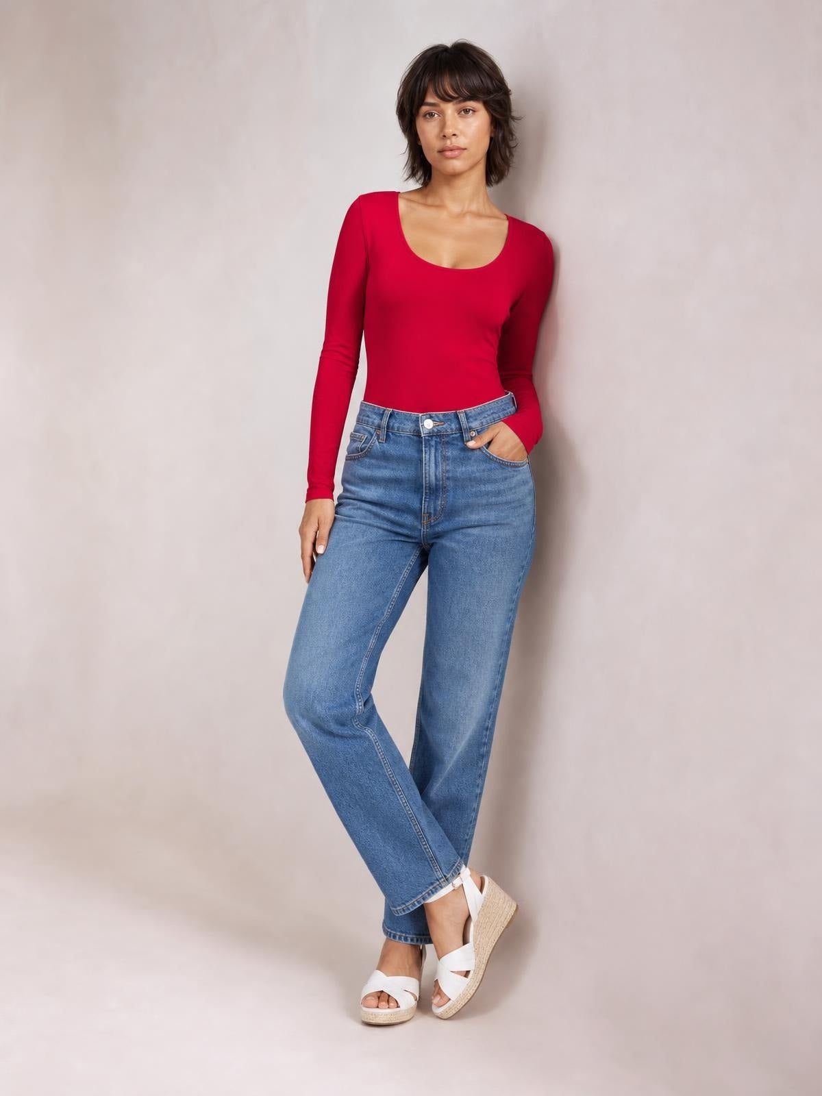 Model wears a fitted red long-sleeve bodysuit with a smooth silhouette, posing with one hand in her jeans pocket and crossed legs.