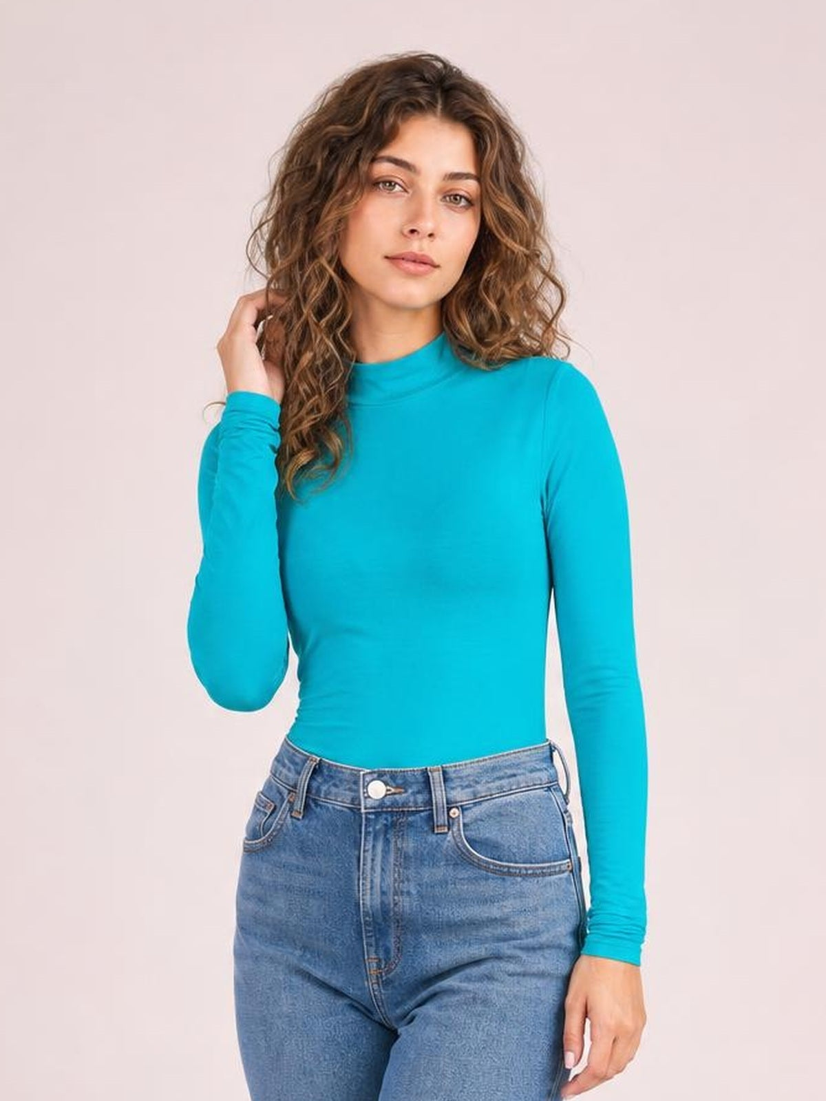 Model poses in a bright turquoise mock neck long sleeve bodysuit with a fitted silhouette, smooth construction, and one hand touching her hair.