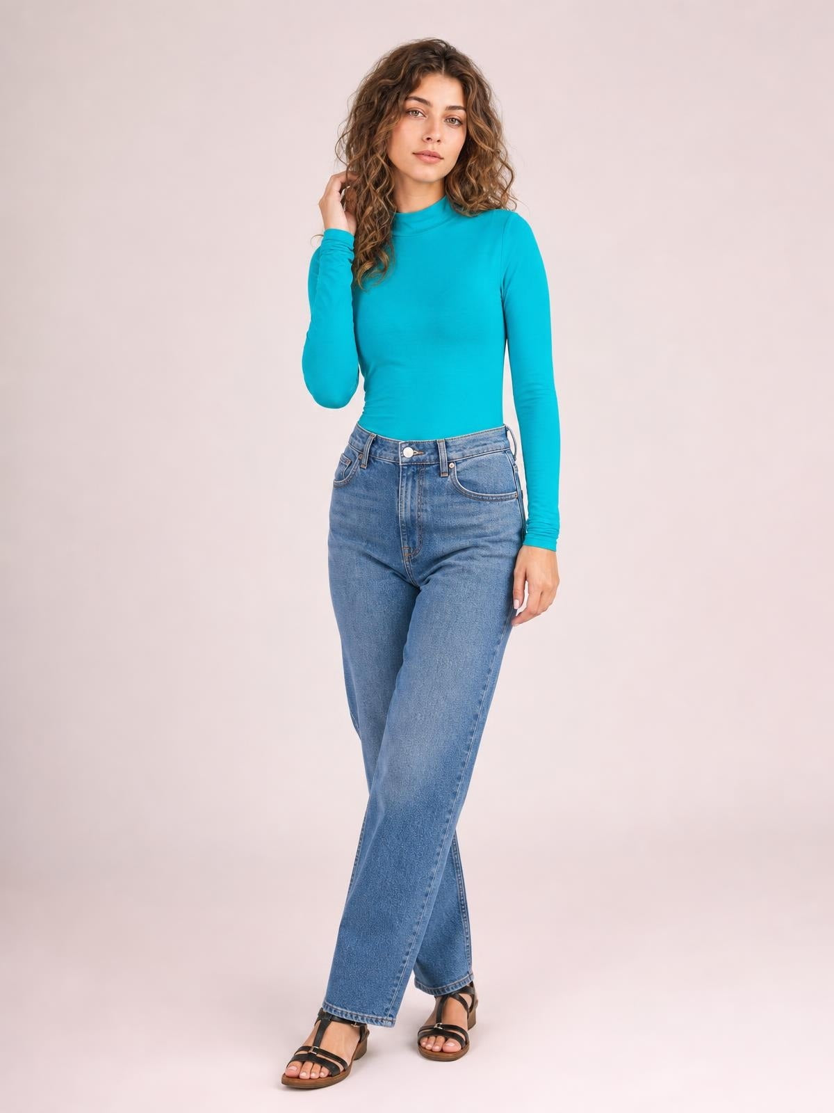 Model in a turquoise mock neck long sleeve bodysuit with a fitted silhouette and smooth opaque construction, posing with one hand in her hair.