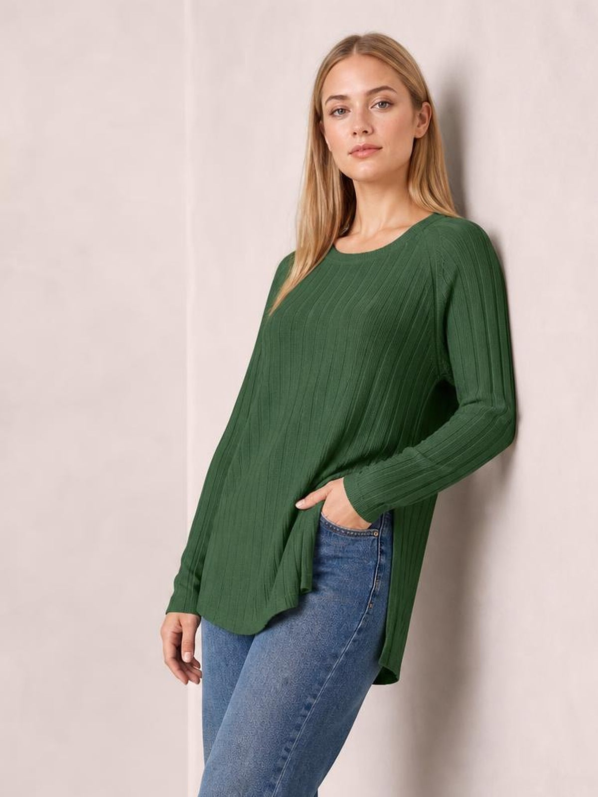 Model poses in a forest green ribbed round neck T-shirt with long sleeves, a relaxed silhouette, and side slits over blue jeans.