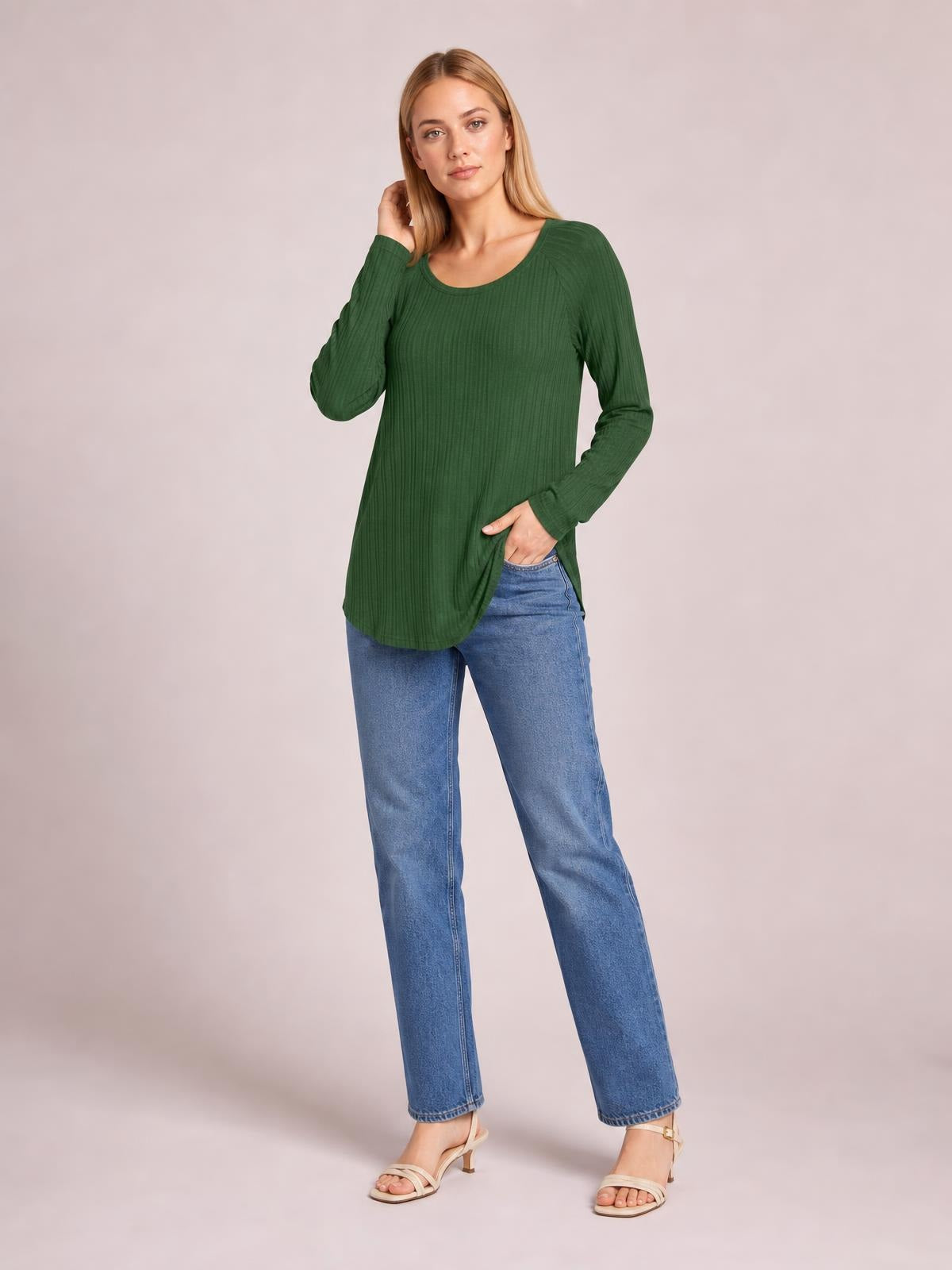 A model poses in a forest green ribbed round-neck T-shirt with long sleeves, a relaxed tunic silhouette, and a visible side slit, gently lifting the hem.