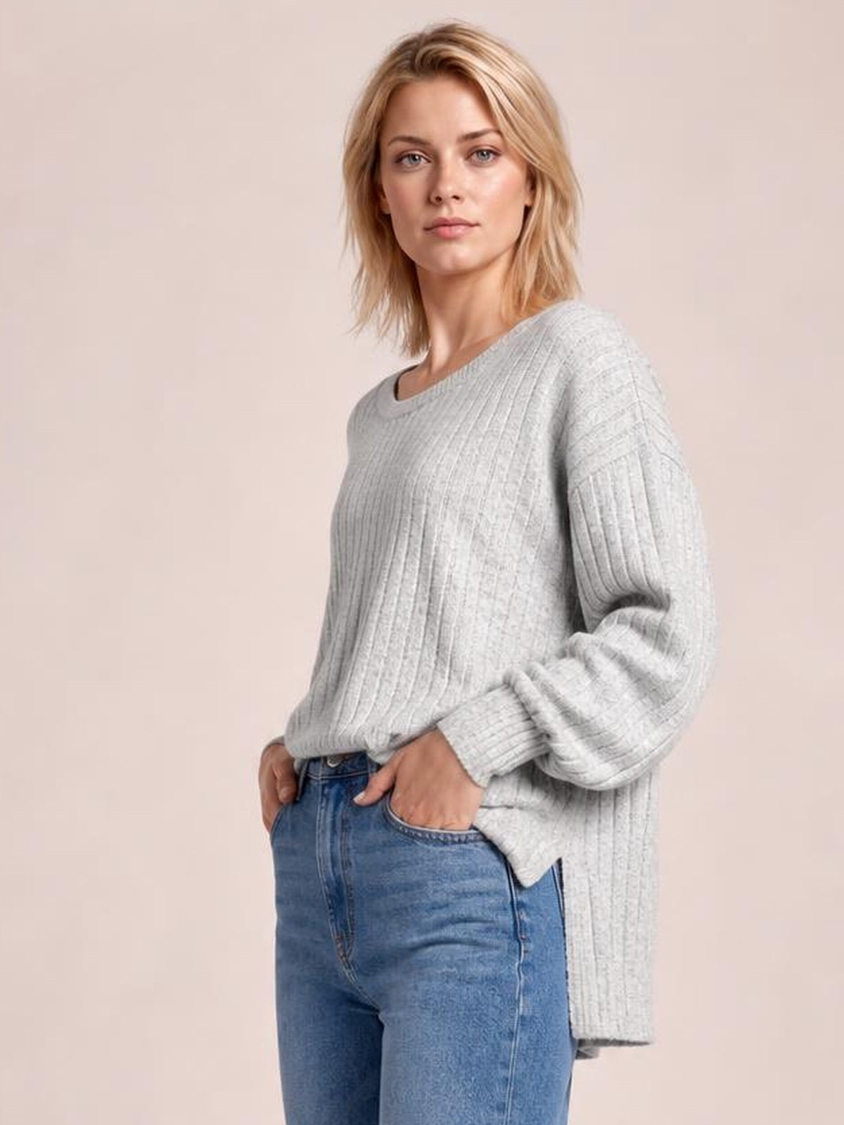 A model wears a light gray ribbed round neck slit T-shirt with long sleeves, a relaxed tunic silhouette, and hands resting in her jeans pockets.