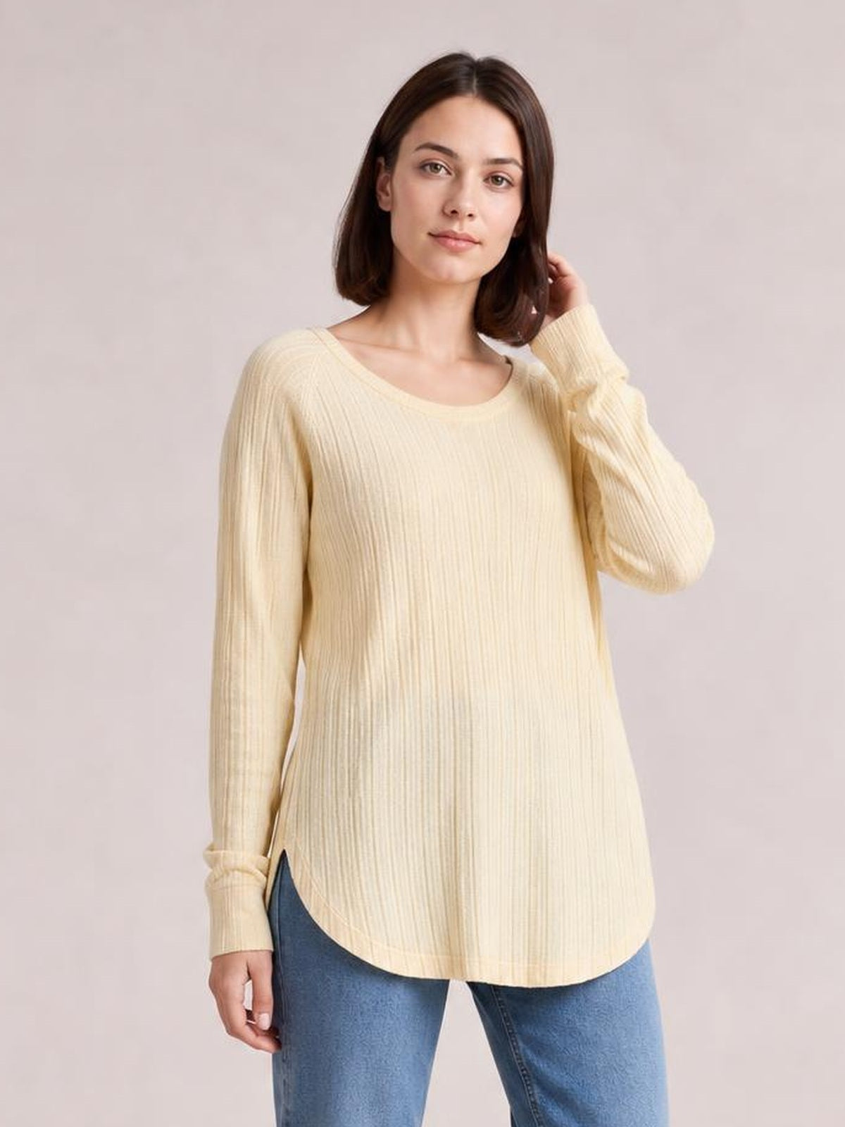 Model wearing a cream ribbed round neck T-shirt with long sleeves, a relaxed silhouette, and curved hem with side slits, touching her hair.