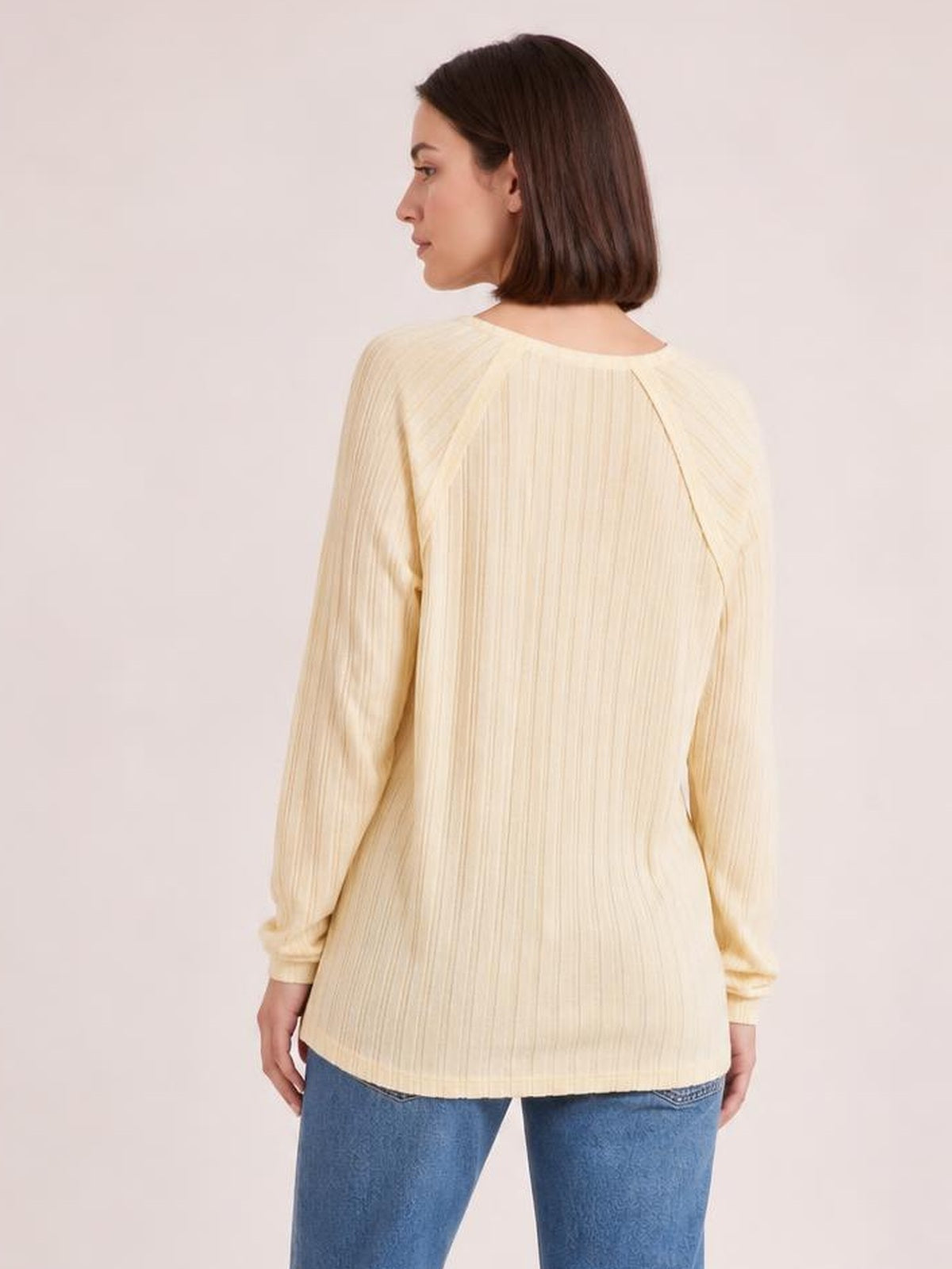 A model looks over her shoulder while wearing a cream ribbed round-neck T-shirt with long sleeves and a relaxed, straight silhouette.