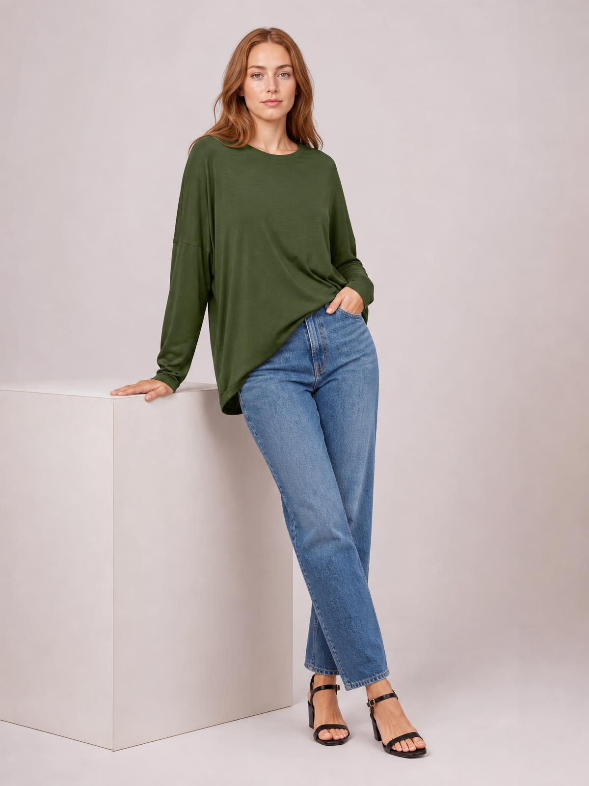 A model wears an olive green round neck dropped shoulder T-shirt with long sleeves and a relaxed silhouette, leaning with one hand on a pedestal.