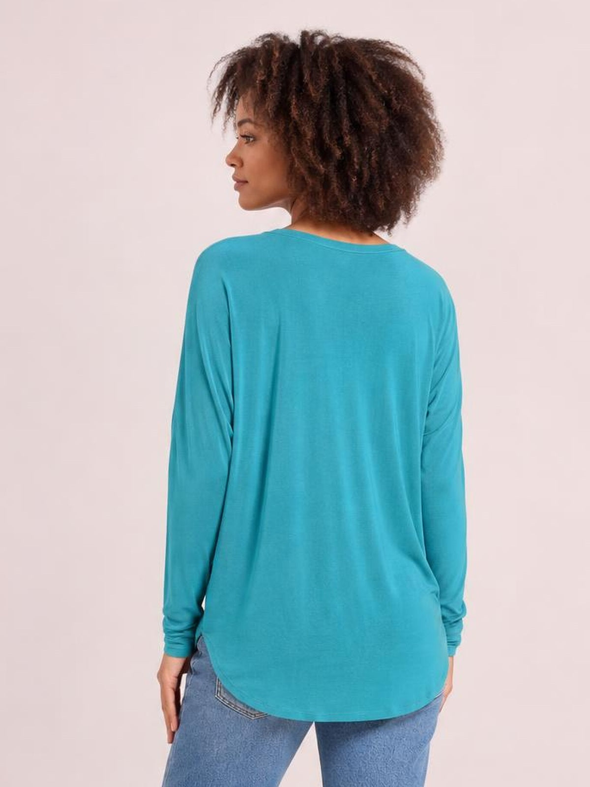 A model turns slightly to show the back of a turquoise round neck dropped shoulder T-shirt with long sleeves, a relaxed silhouette, and curved hem.