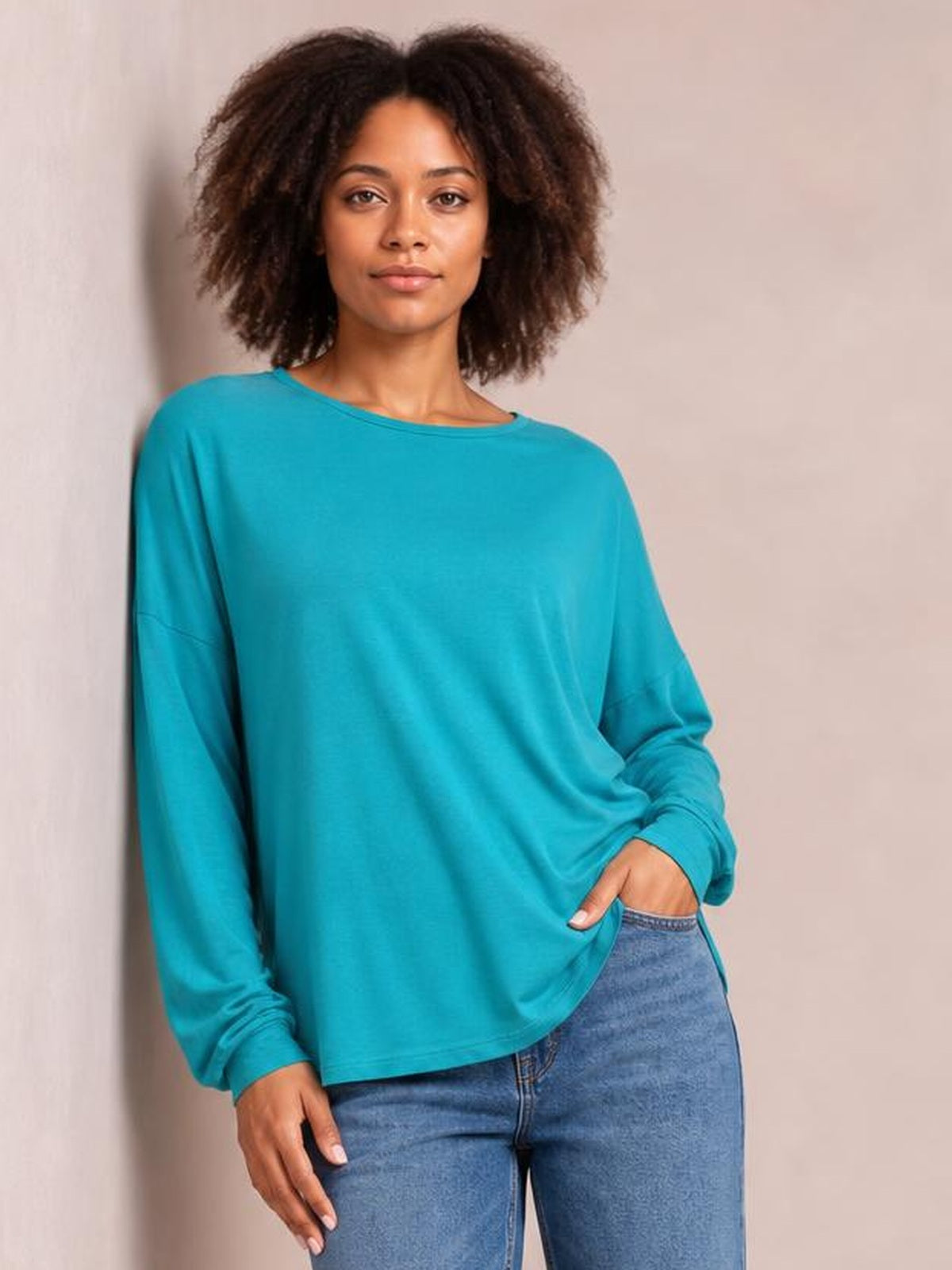 Model wears a turquoise round neck dropped shoulder T-shirt with a relaxed silhouette, long sleeves, and ribbed cuffs while posing with one hand at her waistband.