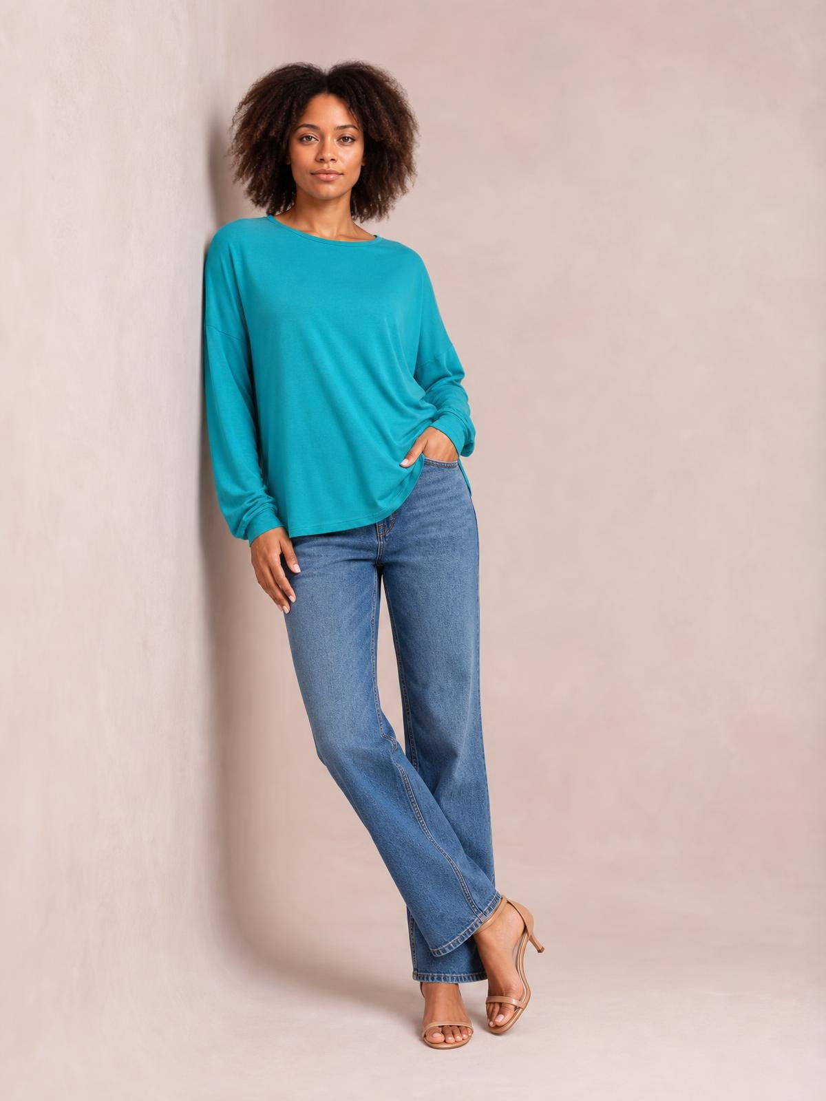 Model wears a turquoise round neck dropped shoulder T-shirt with a relaxed silhouette and long sleeves, standing with one hand in her jeans pocket.