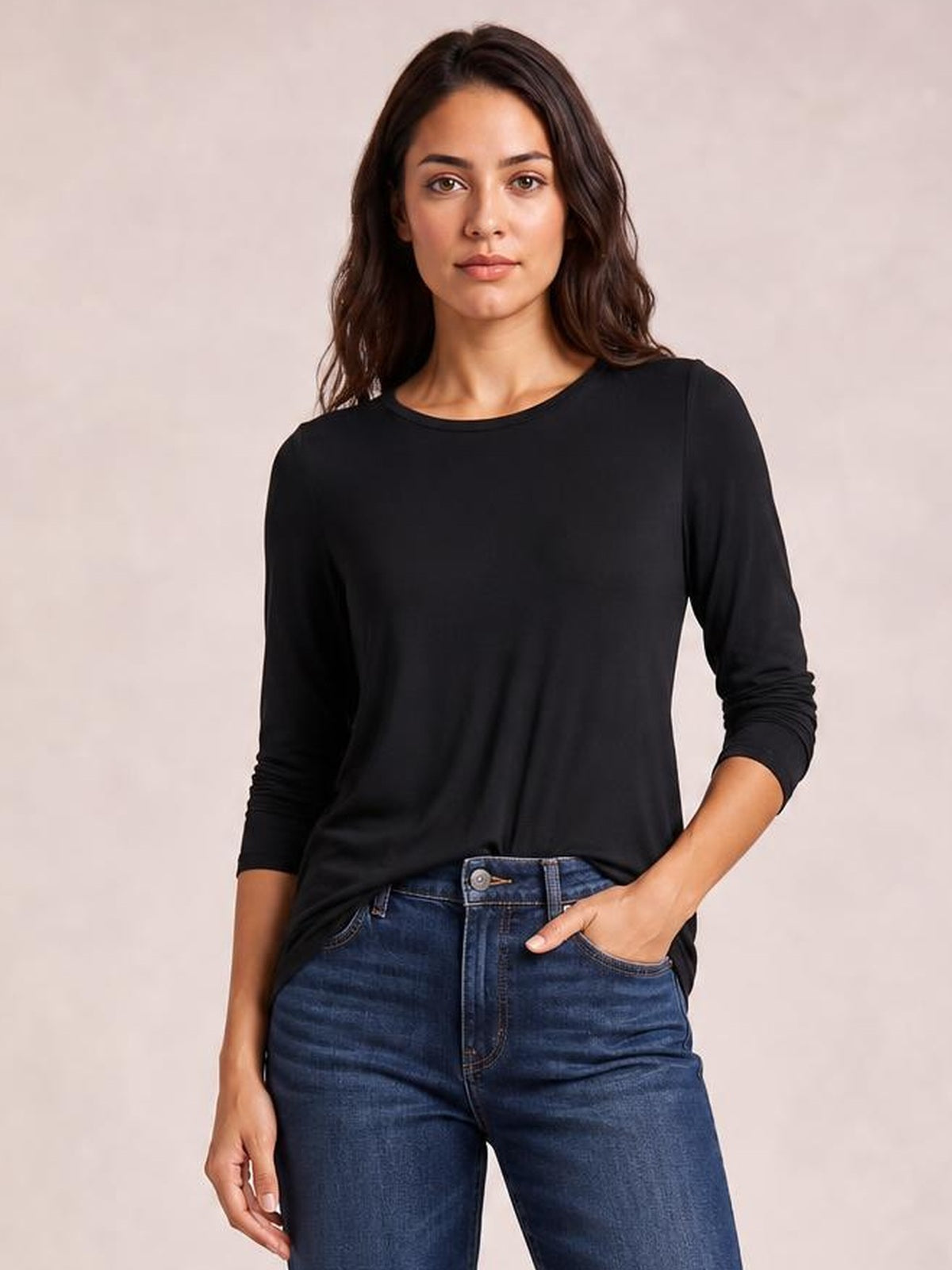 Model wearing a black round neck long sleeve top with a relaxed silhouette and subtle draping, paired with jeans and one hand in a pocket.