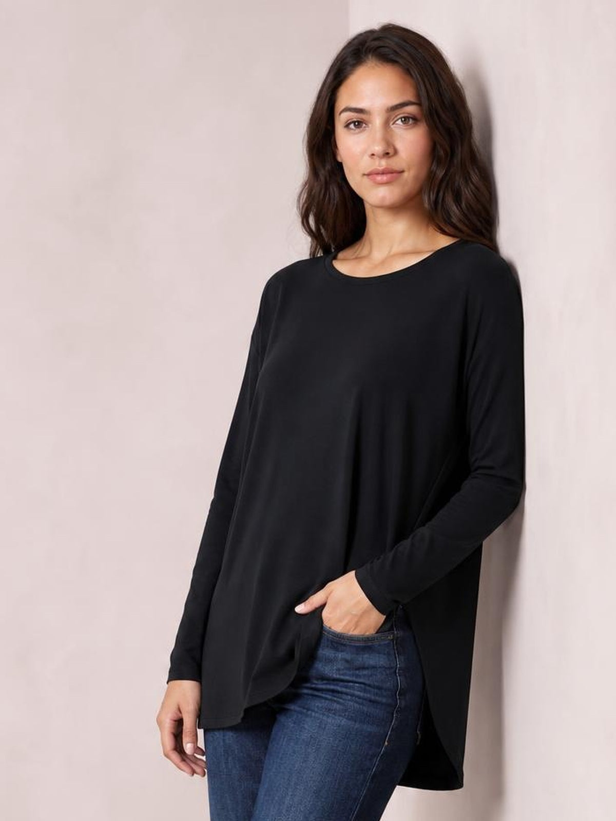 A model stands in a black round neck long sleeve top with a relaxed silhouette, soft drape, and curved high-low hem over blue jeans.