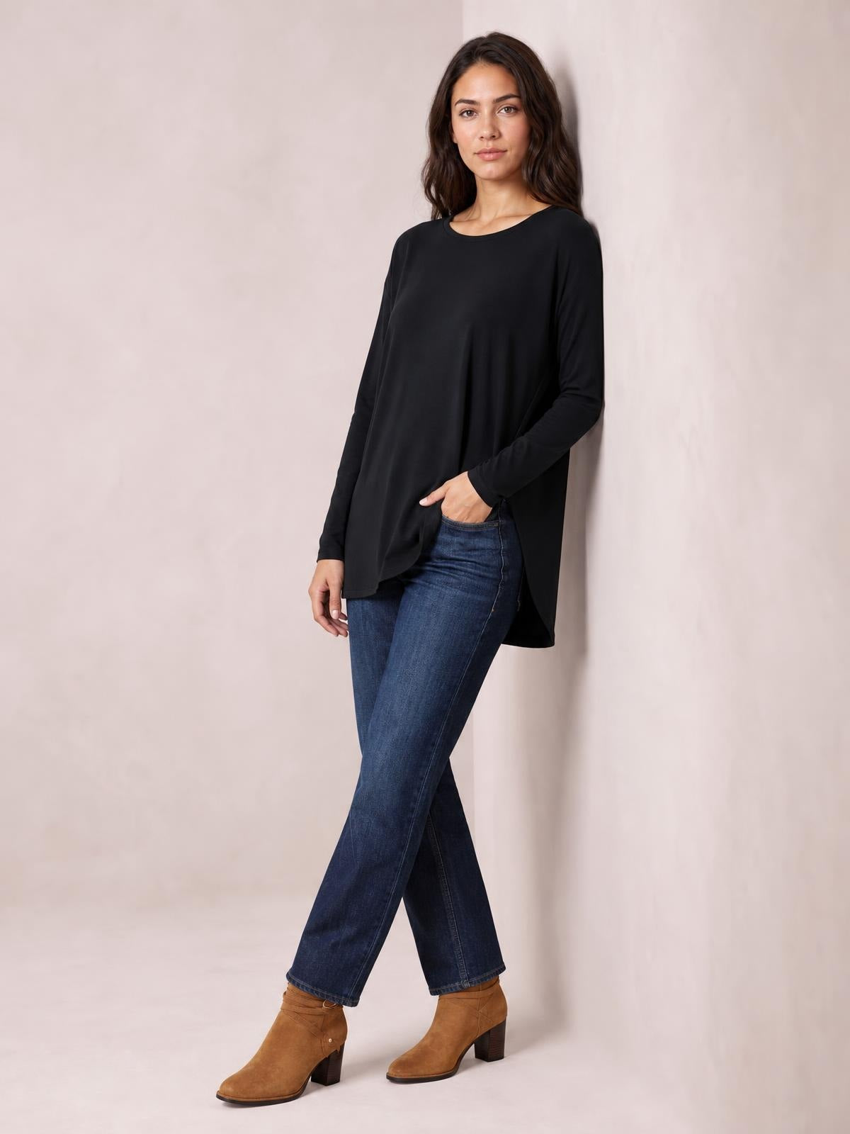 A model poses in a black round-neck long-sleeve top with a relaxed, draped silhouette and longer curved hem, styled with jeans.