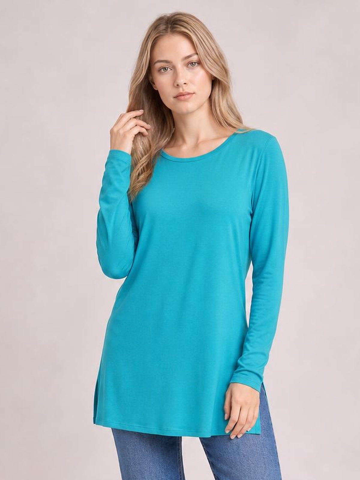 Model poses in a turquoise round-neck long-sleeve top with a relaxed, slightly elongated silhouette and straight hem, lifting one hand to her hair.